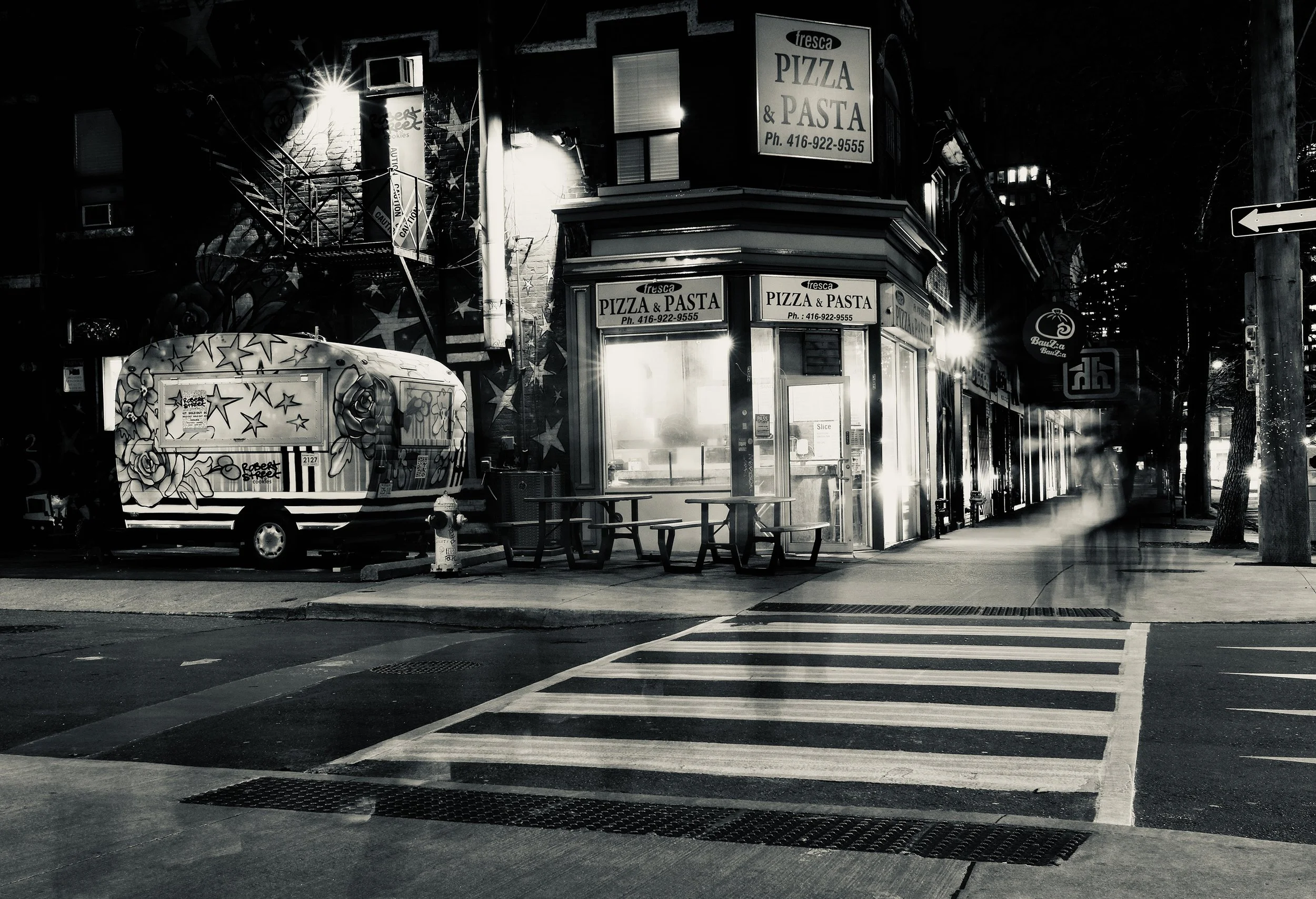 A nighttime street scene with a pizza and pasta shop called 'Fresca Pizza' with illuminated signs, an ice cream truck decorated with stars and flowers, and a food stand, with a crosswalk and a power pole with street signs on the sidewalk. Toronto