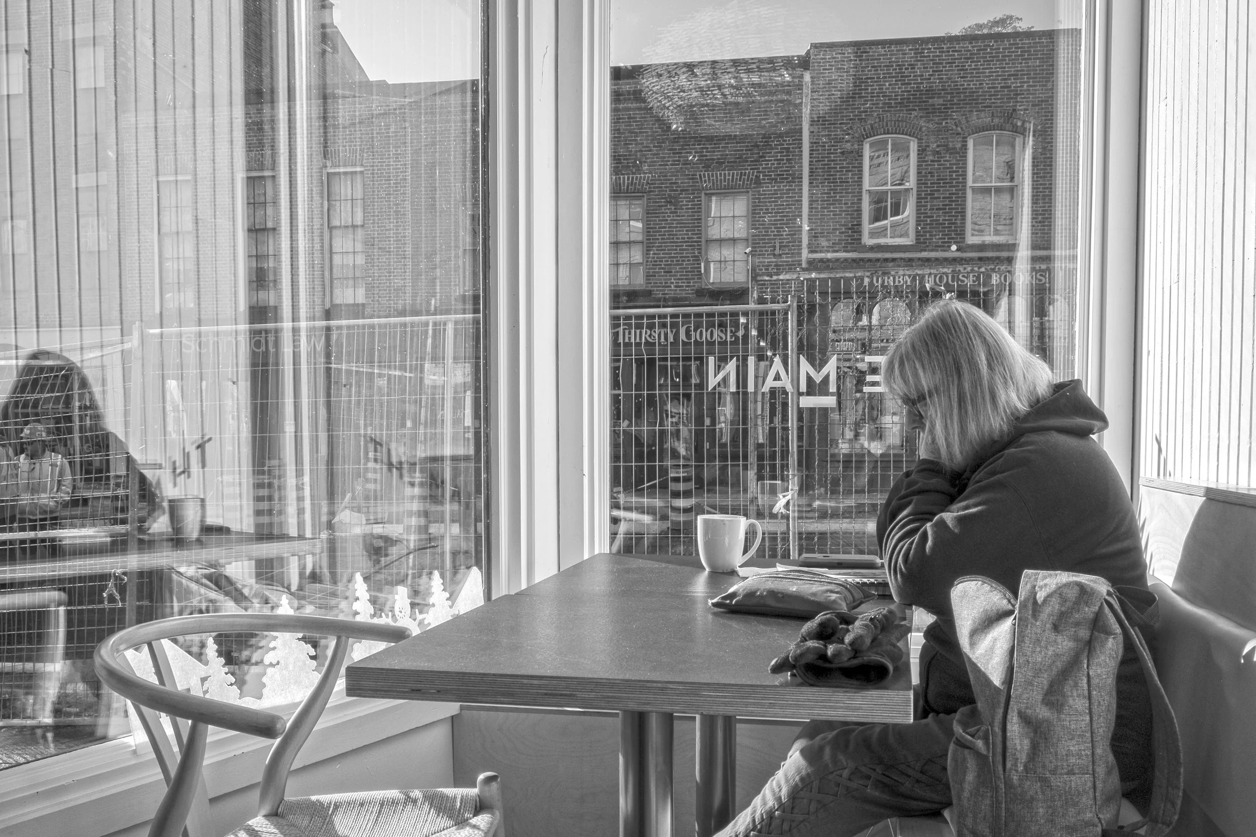 Black and white image of woman sitting at a table in a café window flooded with sunlight.