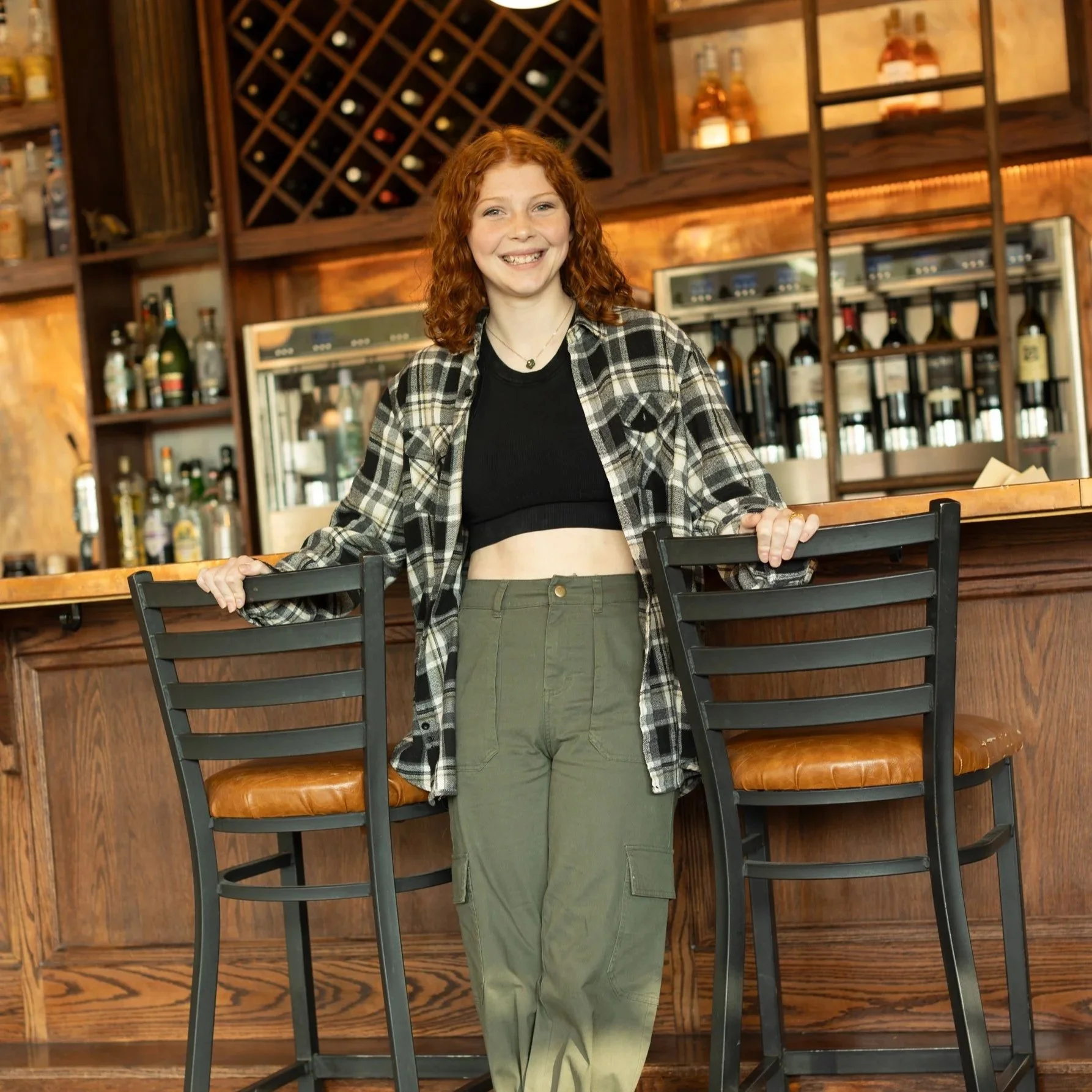 A young woman with red curly hair smiling at a bar, wearing a black crop top, green cargo pants, and a plaid shirt.