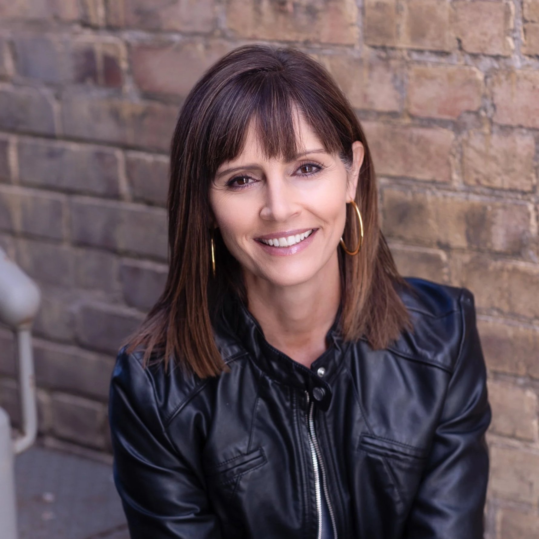 A woman with shoulder-length brown hair, wearing gold hoop earrings and a black leather jacket, smiling in front of a brick wall.