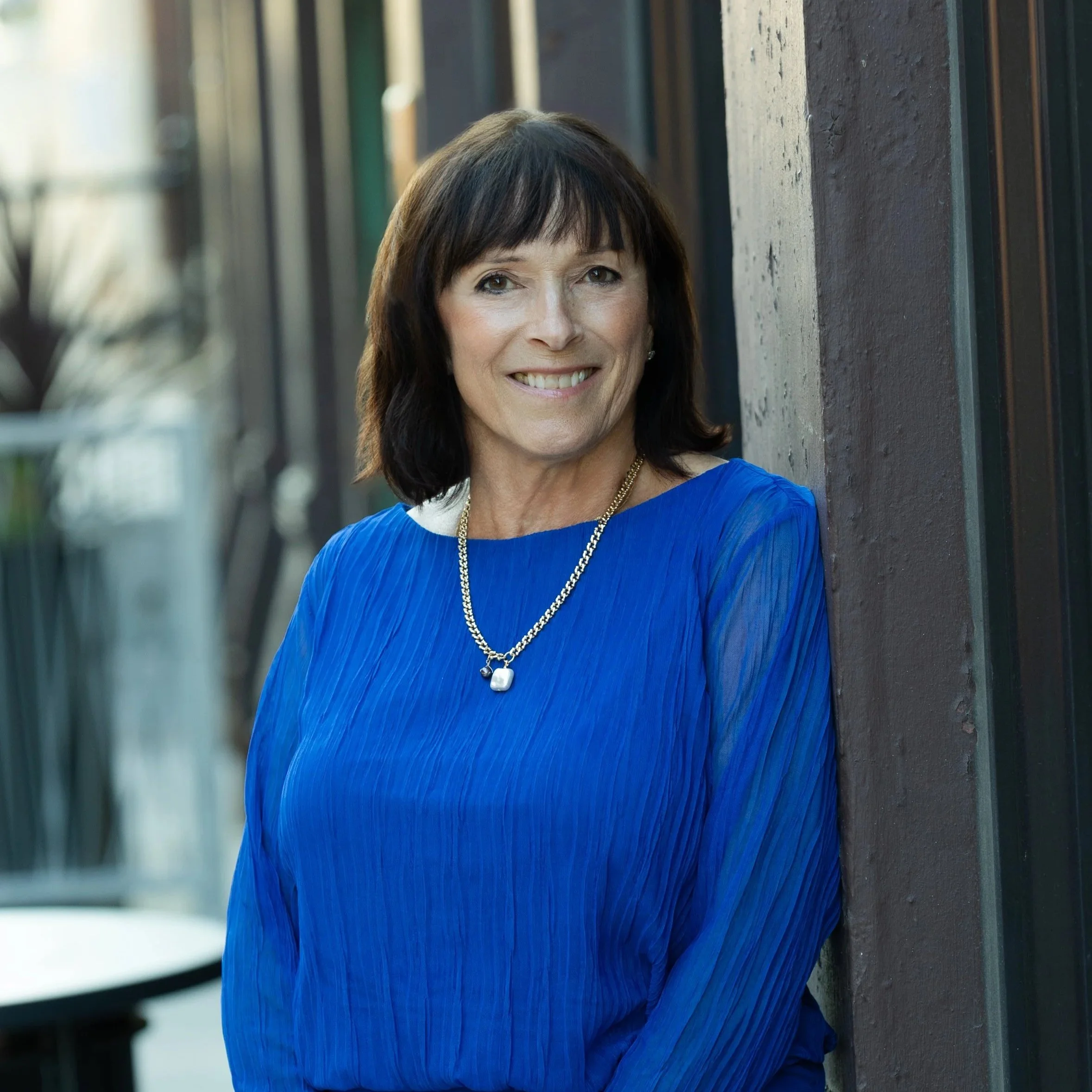 A woman with dark brown hair and a bright smile, wearing a vibrant blue dress with sheer sleeves and a gold necklace, standing outdoors against a dark wall with blurred background.