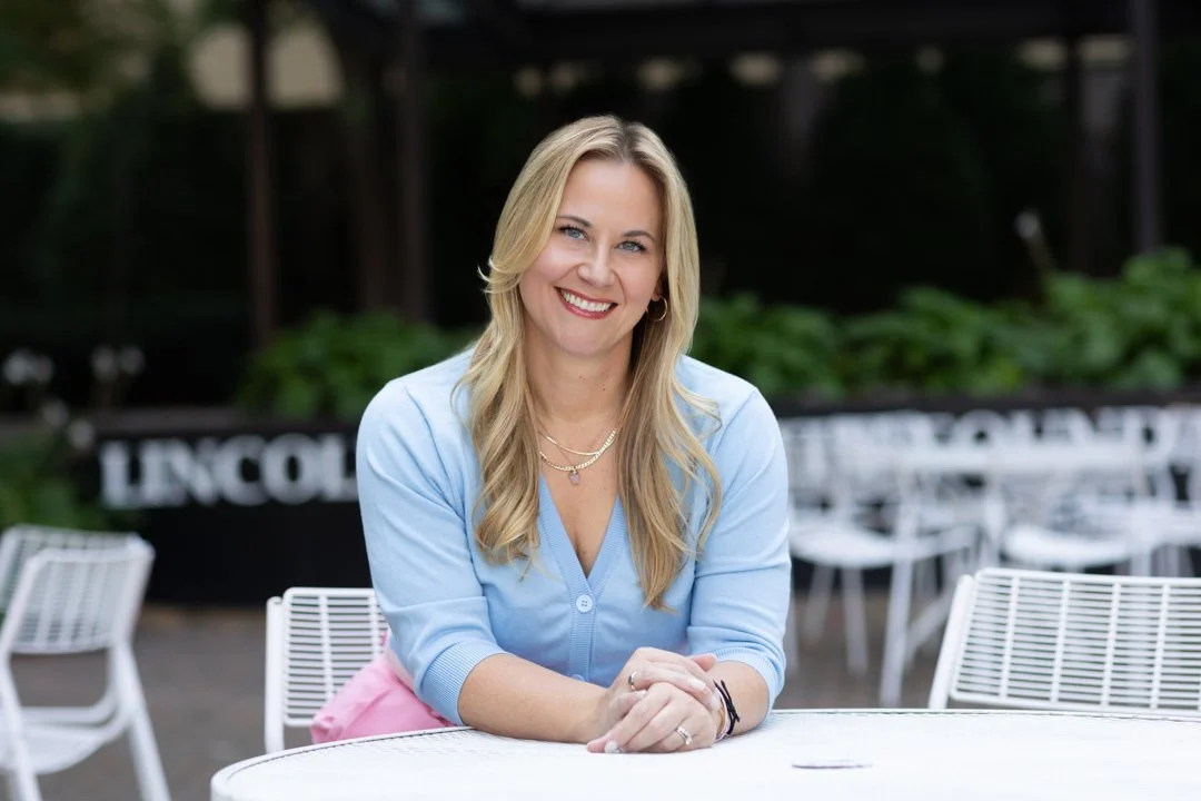 A woman with blonde hair smiling and sitting at a white table outdoors, wearing a light blue top and pink skirt.