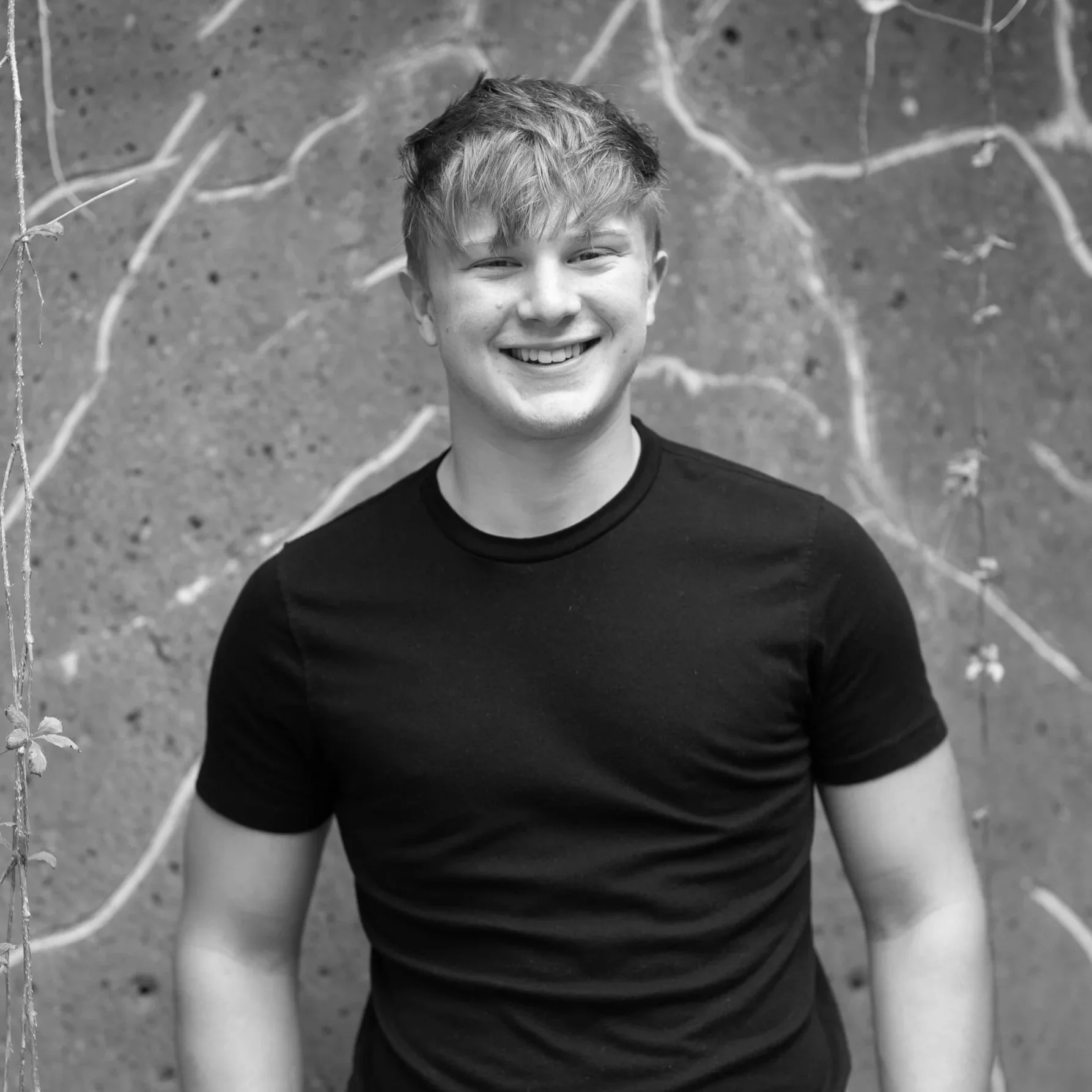 A young man with short, tousled hair smiling, wearing a black t-shirt, standing outdoors against a background of a textured wall with branches.