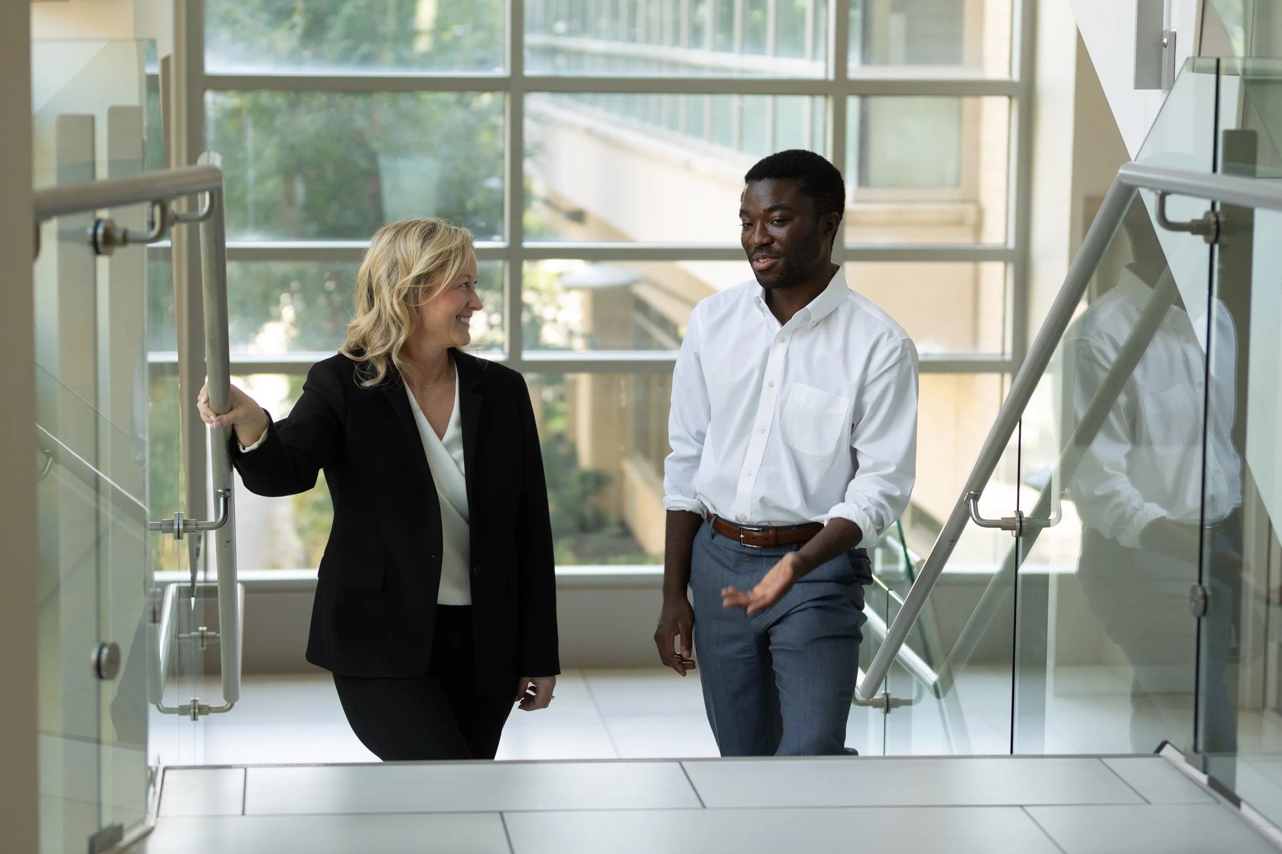 two business professionals talking while walking up stairs.   Windows are behind them.