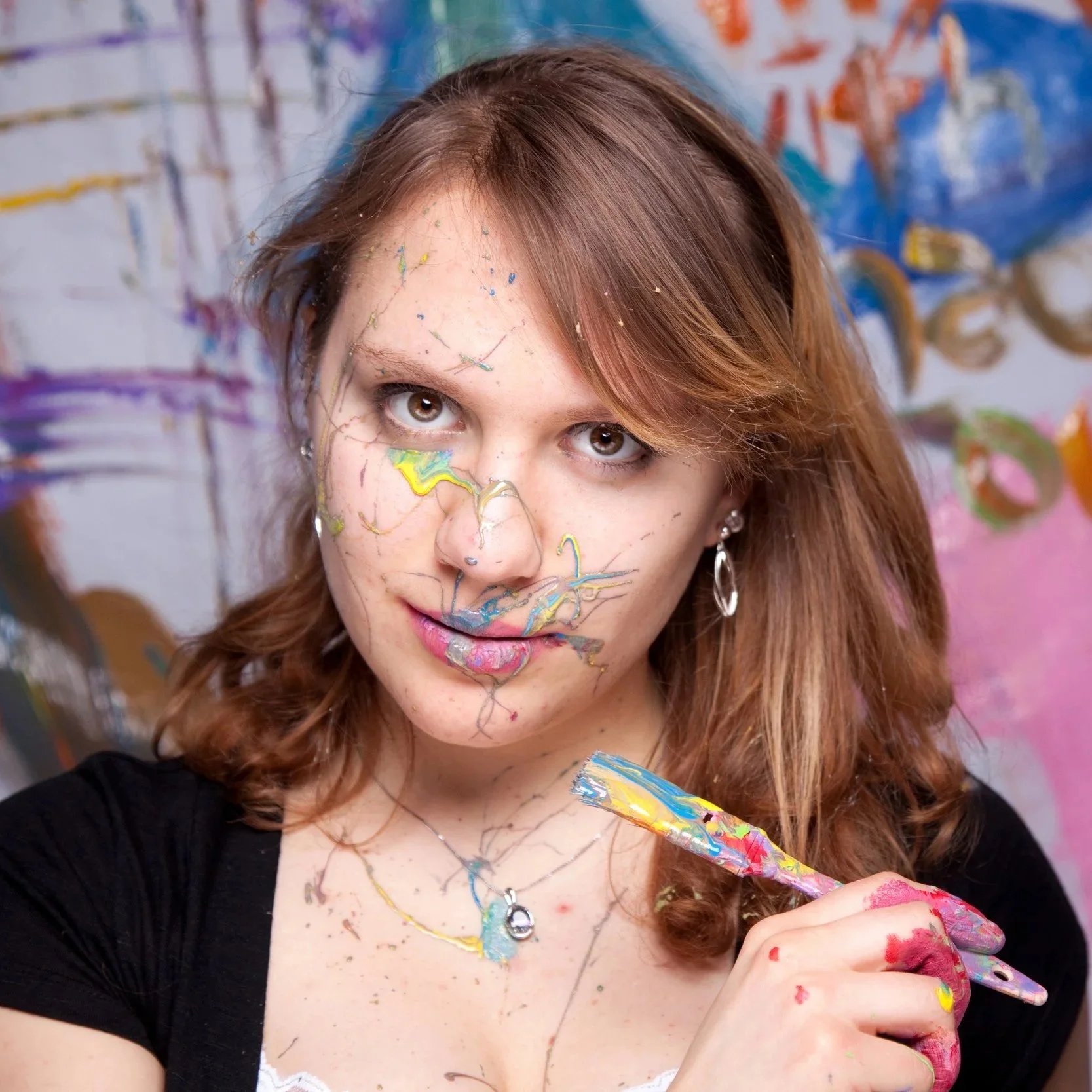 A woman with paint on her face and hands, holding a paintbrush, standing in front of a colorful abstract background.