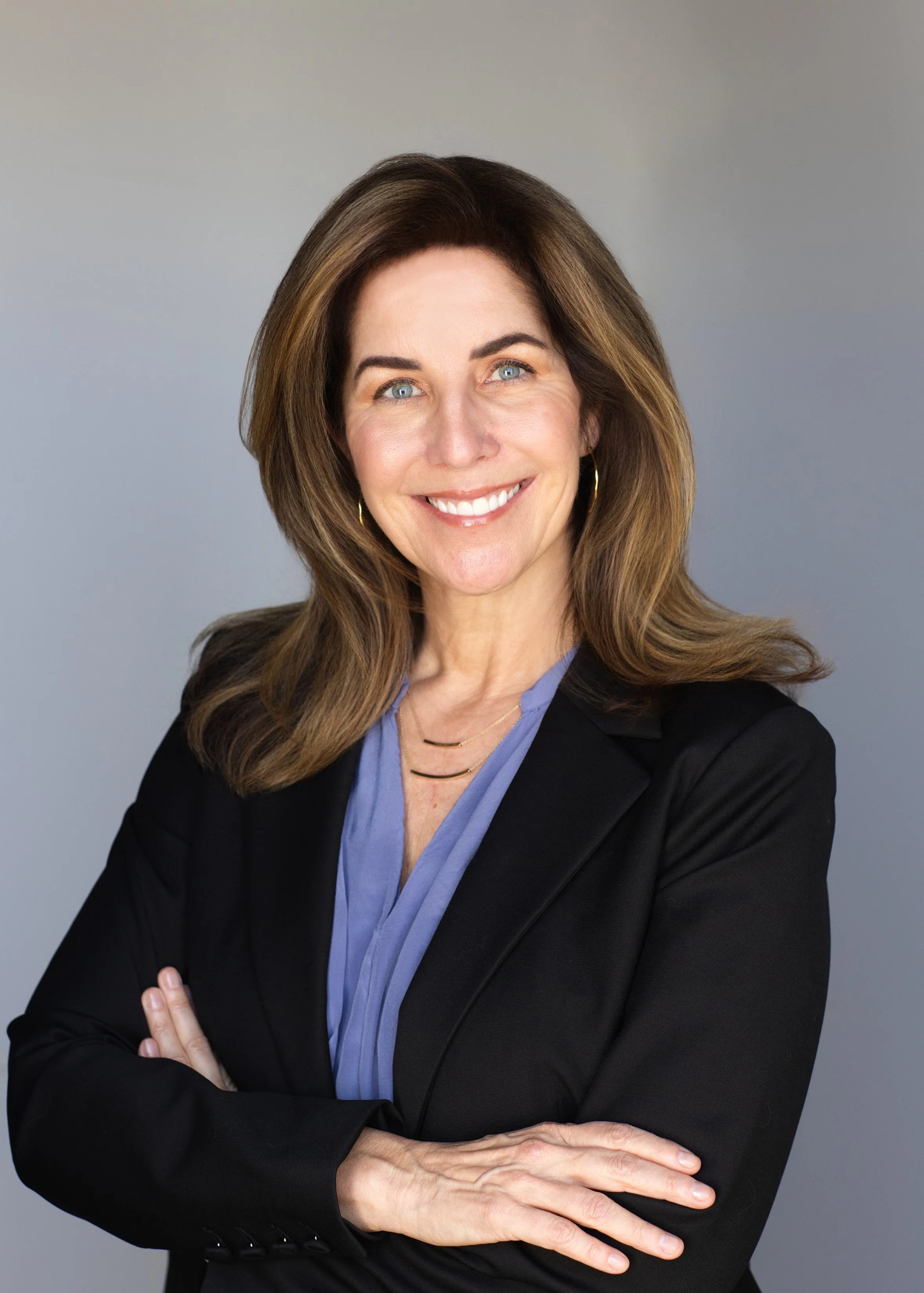 A professional woman with shoulder-length brown hair, blue eyes, and a bright smile, wearing a black blazer, a light blue blouse, gold hoop earrings, and a layered gold necklace, standing with arms crossed against a plain gray background.