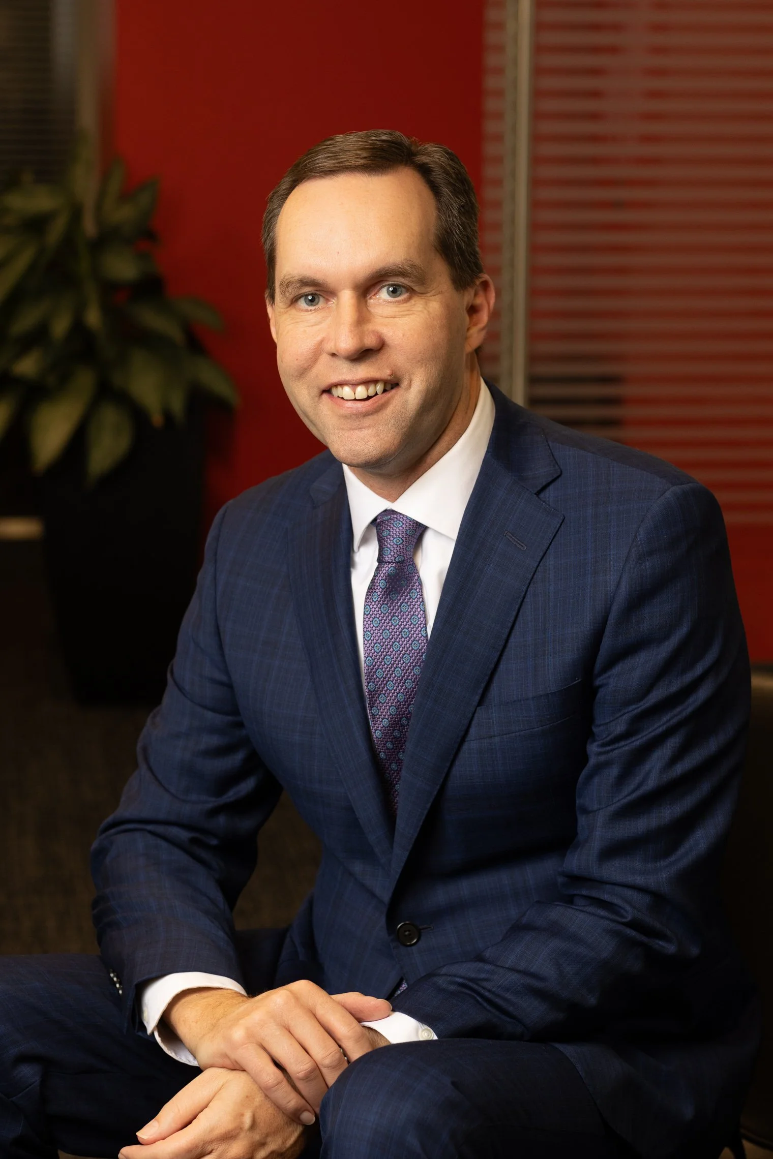 Corporate headshot of a business professional in an office with red walls and blinds.
