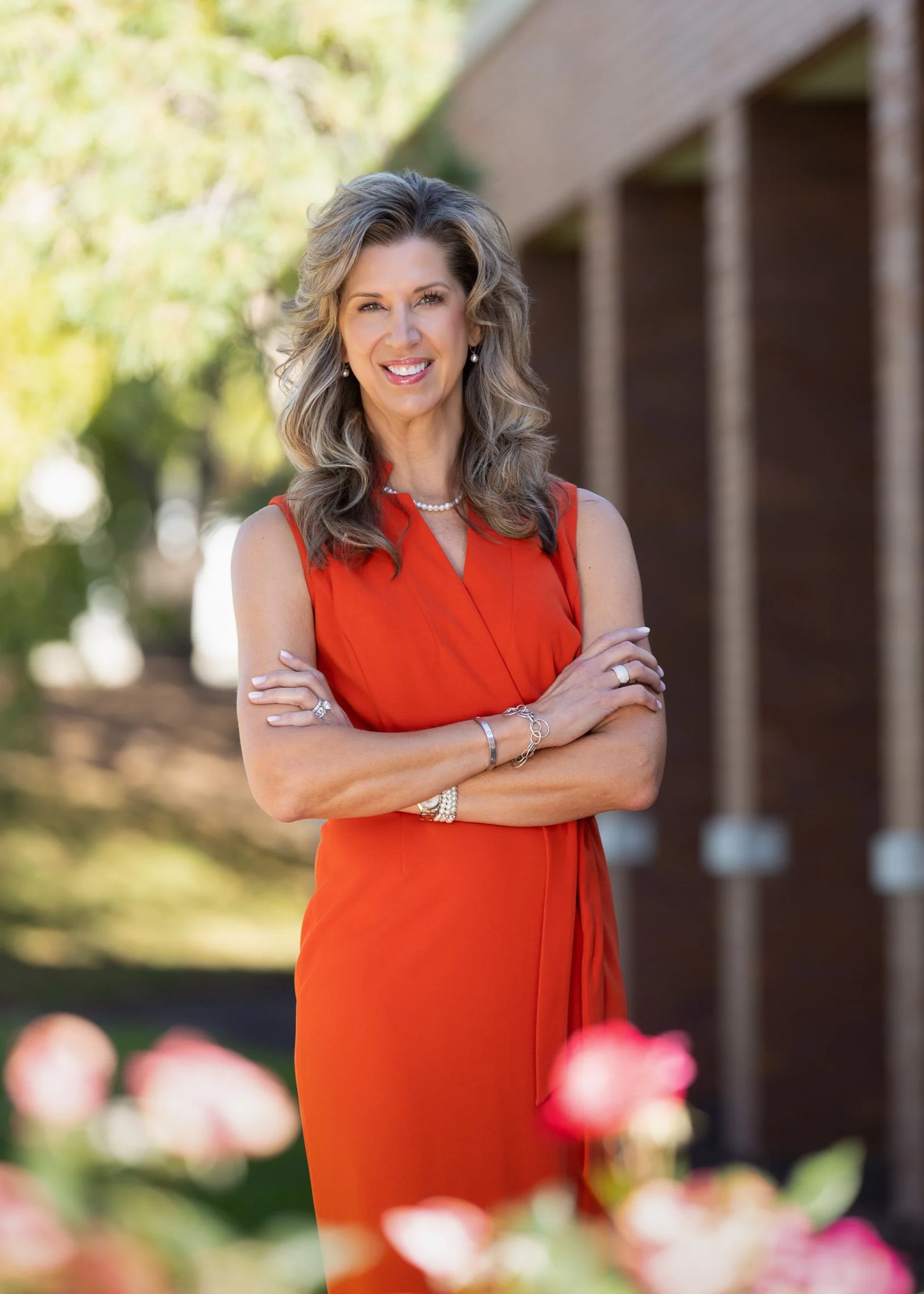 Portrait of a businesswoman in a red dress standing outside.   flowers are in the foreground and building and trees in the background