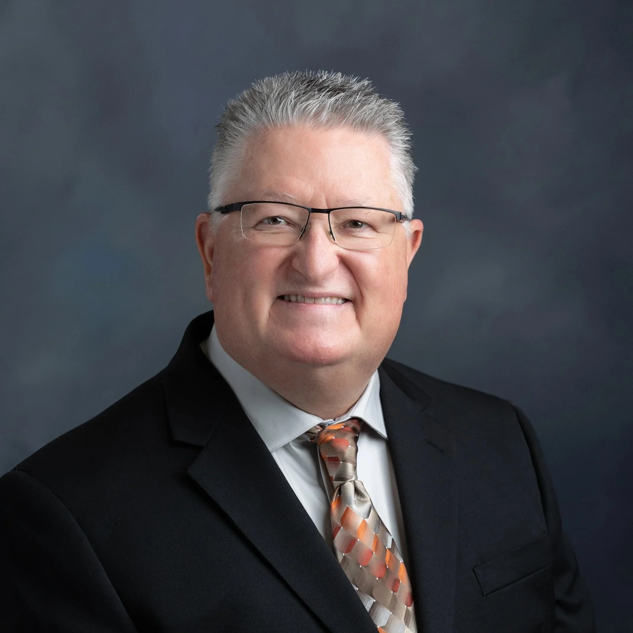 Professional headshot of a middle-aged man with gray hair, glasses, wearing a black suit, white shirt, and patterned tie, against a dark background.