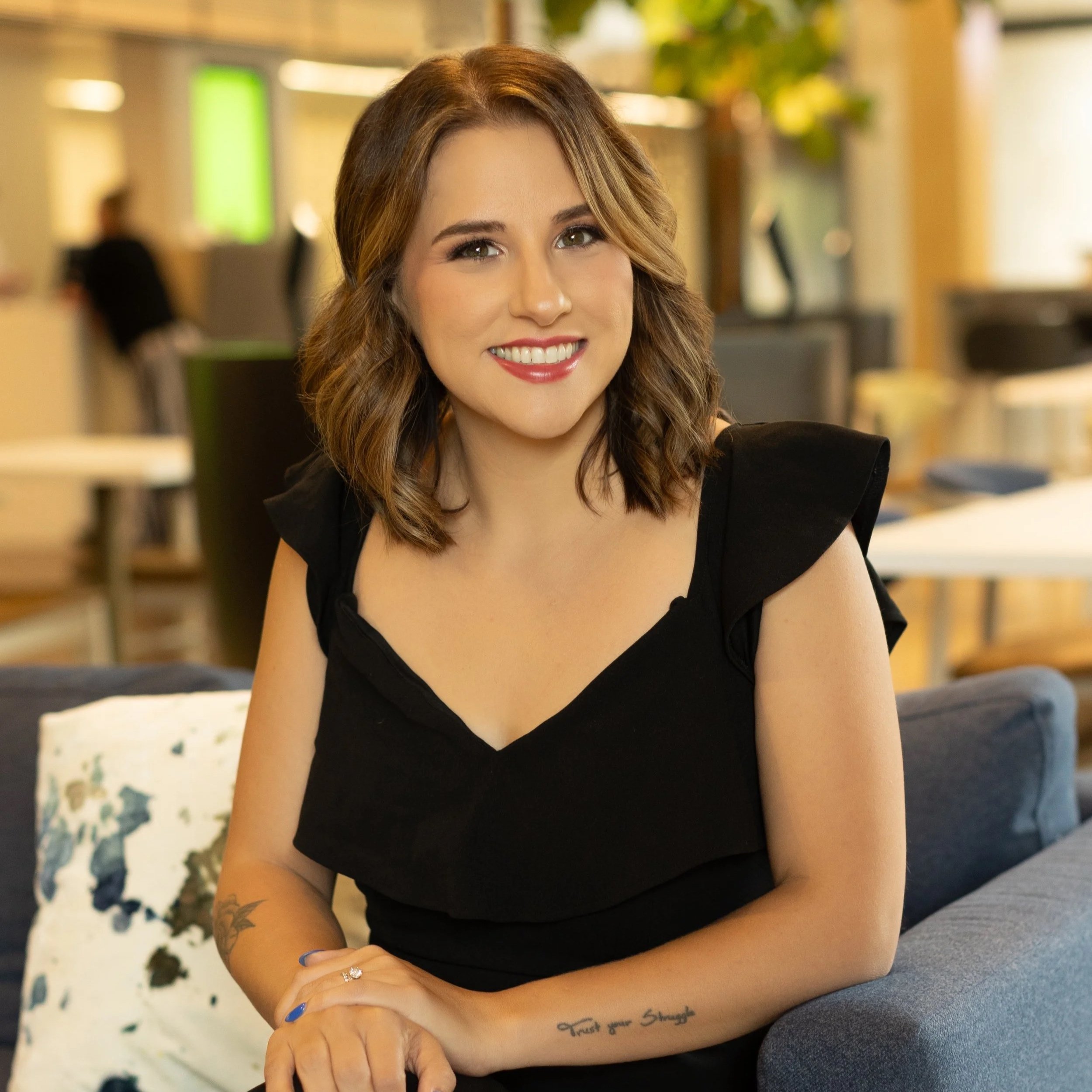 A woman with brown, wavy hair wearing a black top, sitting on a couch in a brightly lit indoor space.