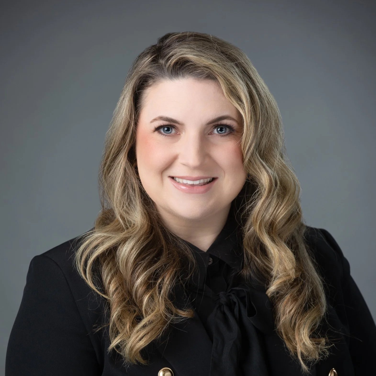 Professional headshot of a woman with long, wavy blonde hair, blue eyes, wearing a black blazer and a black blouse with a bow tie, smiling against a gray background.