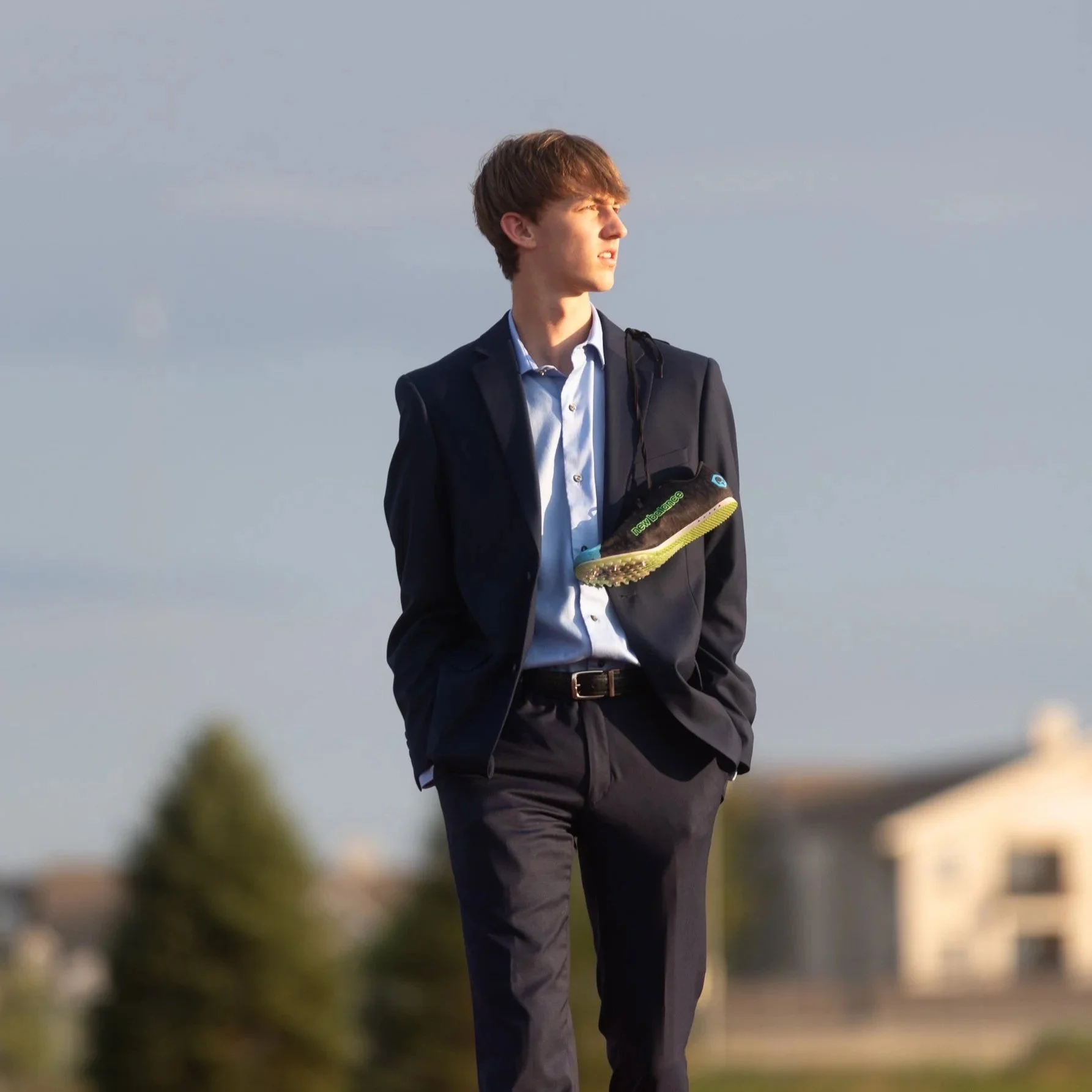 A young man in a dark suit with a blue shirt, standing outdoors with his hands in his pockets, looking into the distance. A black sneaker with green accents hangs from his neck by the shoelaces.