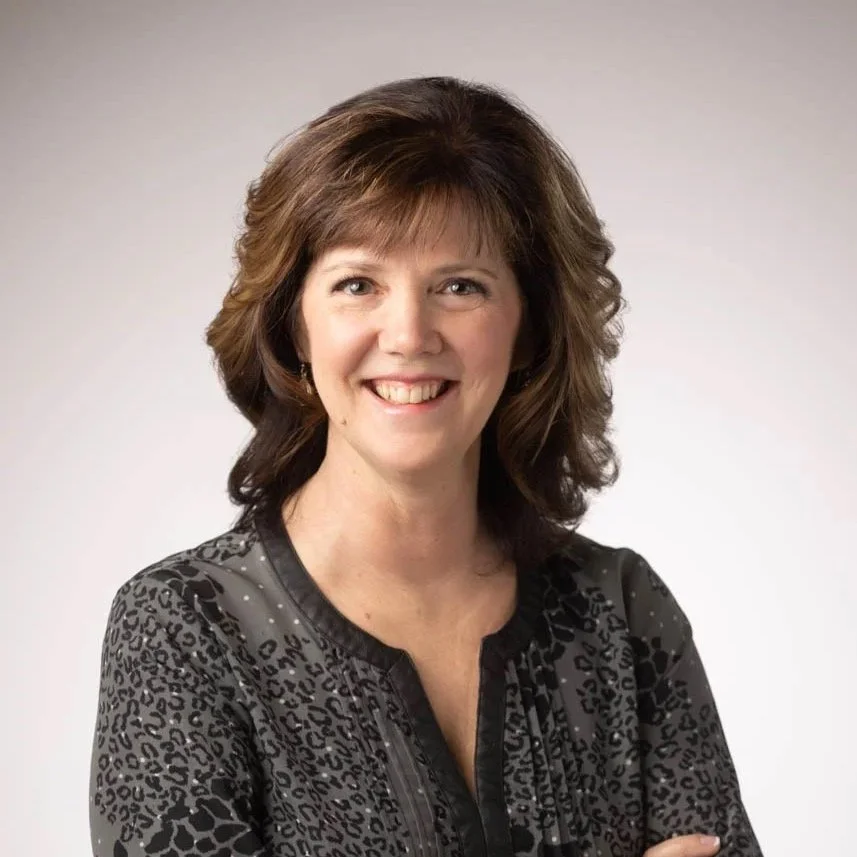 A smiling woman with short, wavy brown hair wearing a patterned dark blouse, posing against a plain light background.
