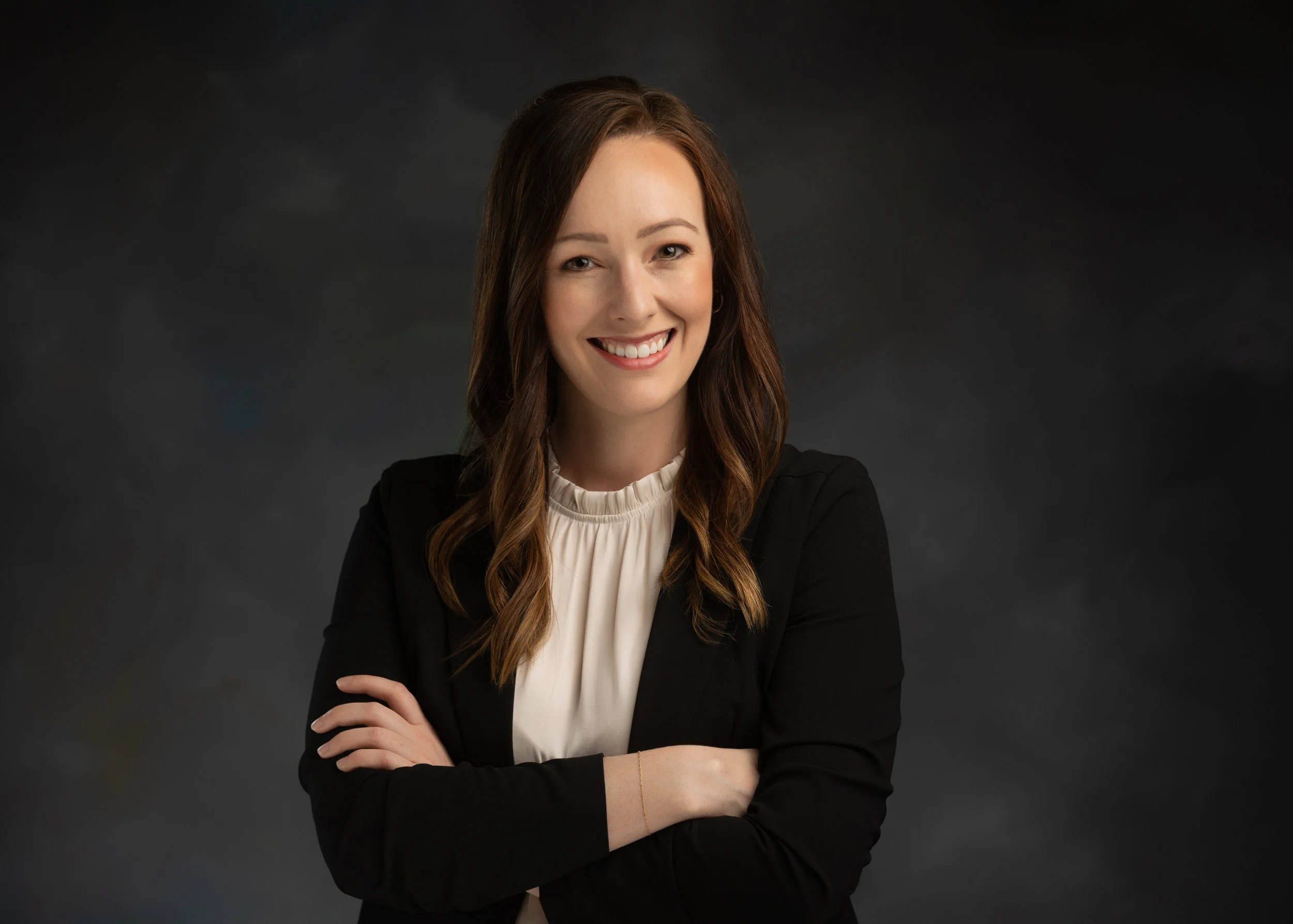 Portrait of a woman with shoulder-length brown hair, wearing a black blazer and a cream-colored blouse, smiling with arms crossed against a dark background.