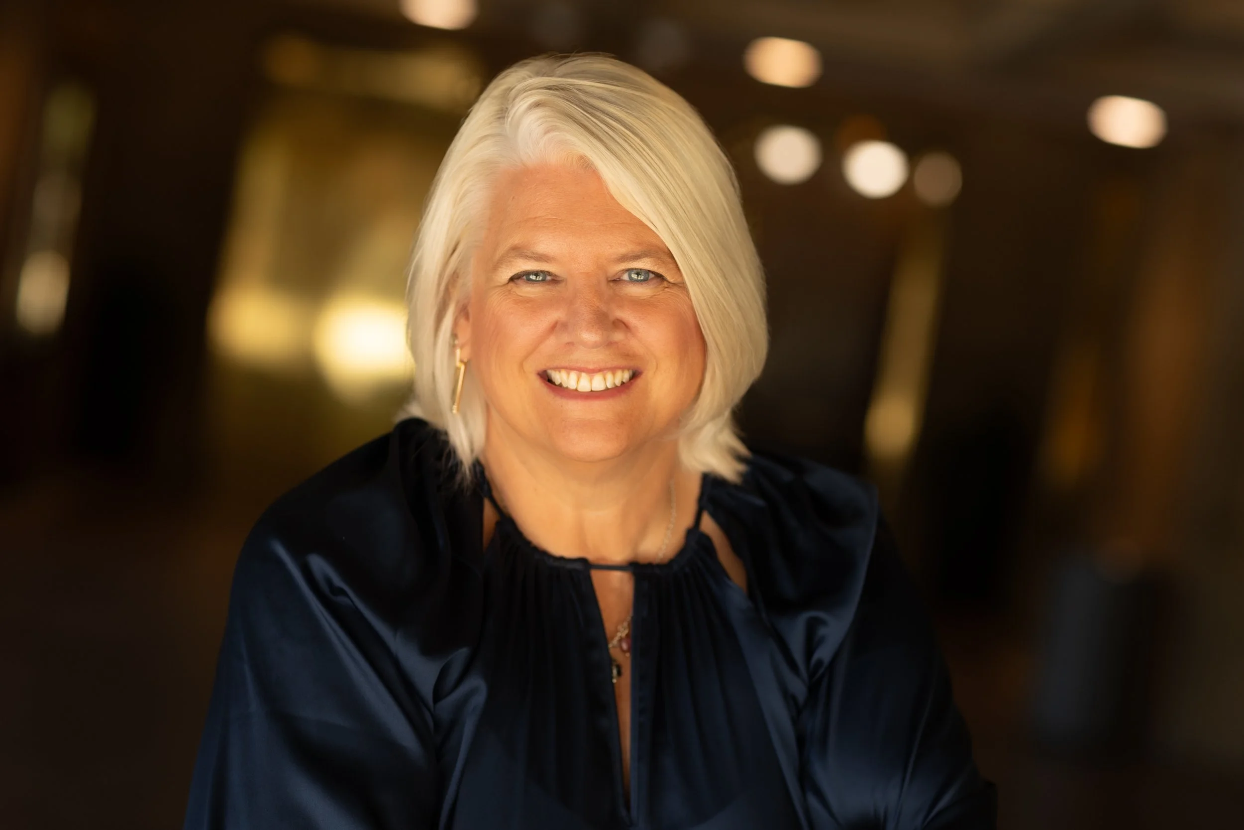 A middle-aged woman with blonde hair and blue eyes, smiling, wearing a dark blue satin blouse with a decorative collar and hoop earrings, in a warmly lit indoor setting.