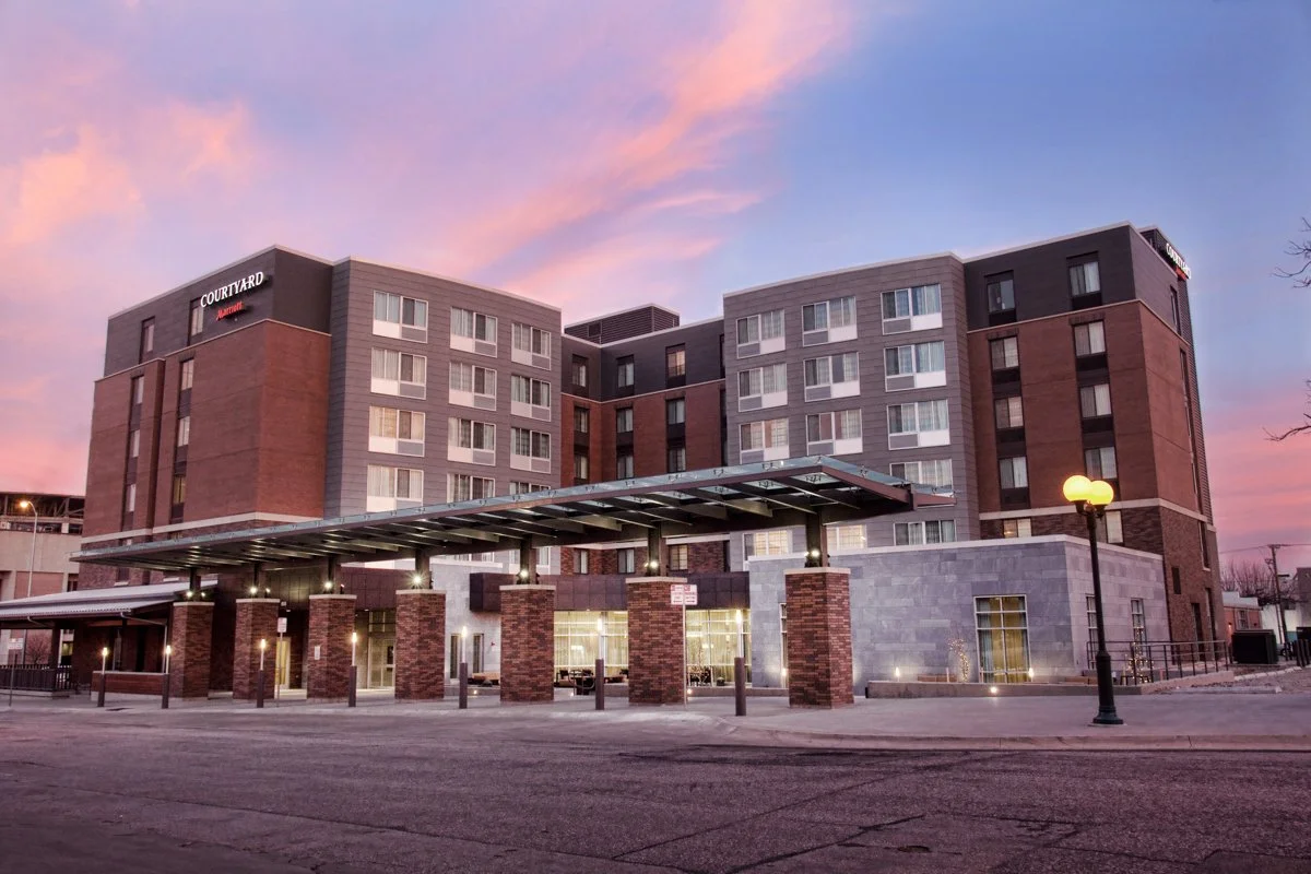 A modern multi-story hotel building with a brick and gray exterior, illuminated by exterior lighting at dusk, with a covered entrance supported by brick columns and a streetlamp in the foreground.