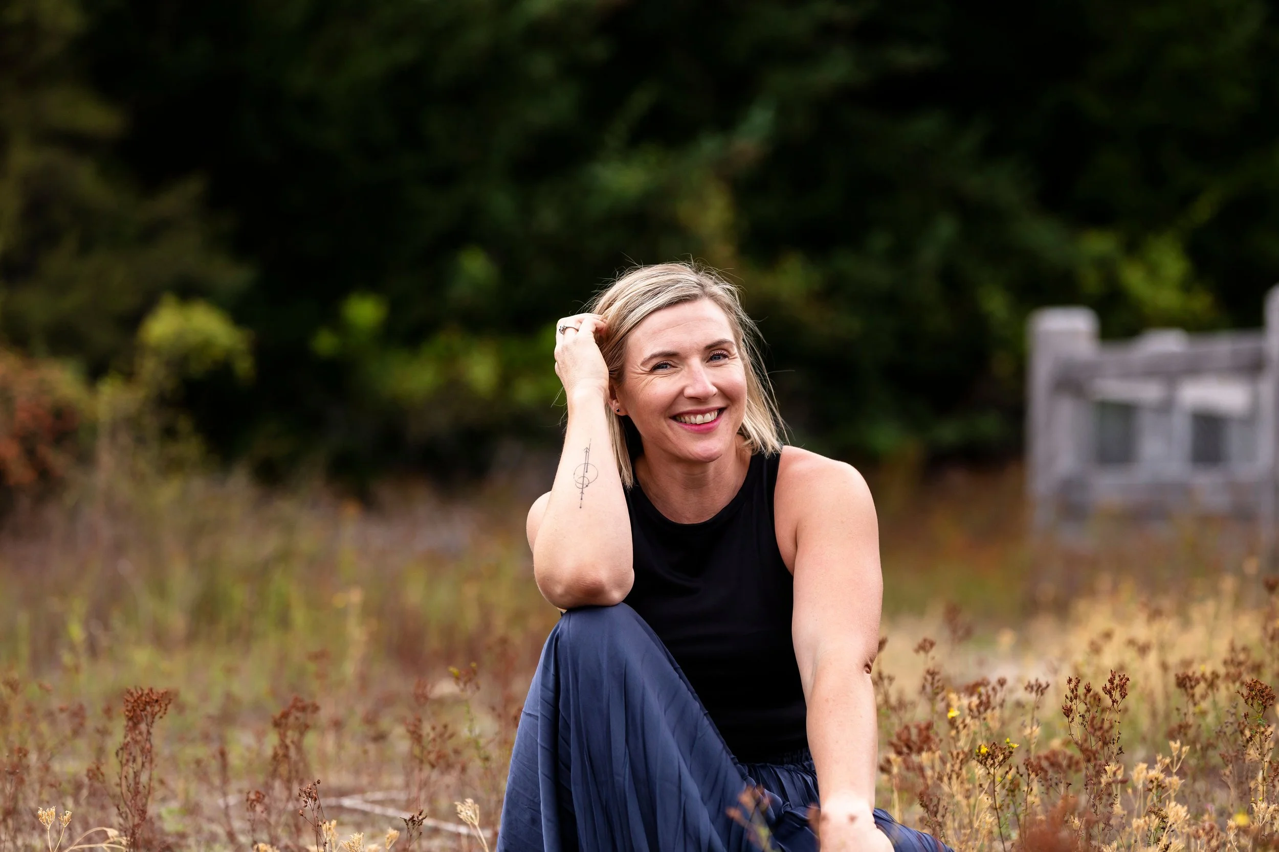 A woman with blonde hair smiling and sitting outdoors in a field of plants during daytime, with a background of green trees and a wooden fence.