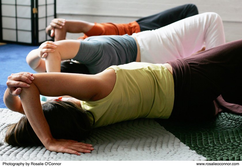 Three people lying on their sides on mats, taking a Feldenkrais group class (A.T.M. - Awareness Through Movement) with knees bent and arms over their heads, in a studio setting.