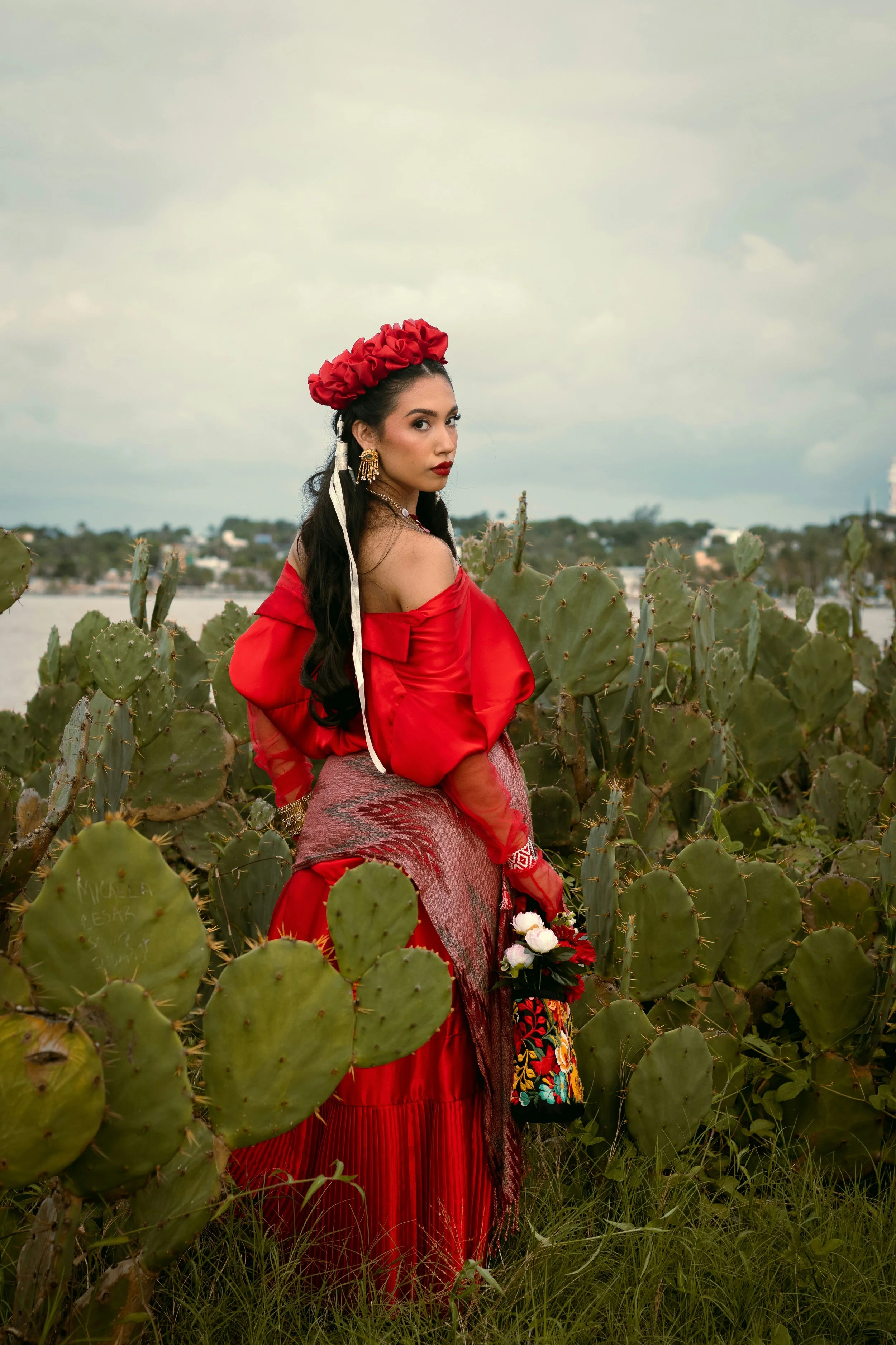 woman with long black hair and darker complexion  wearing a red silk off shoulder dress, a red silk headpiece that looks like flowers carrying a bag that has  bright colored flowers on it with 3 white and light pink roses surrounded by cacti
