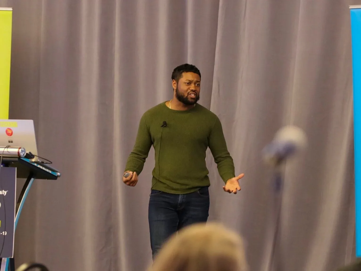Fenton Jagdeo with dark curly hair and a beard wearing a green sweater and jeans, speaking with hand gestures on a stage with a gray curtain backdrop. There are banners on either side of the stage, one yellow and one blue.