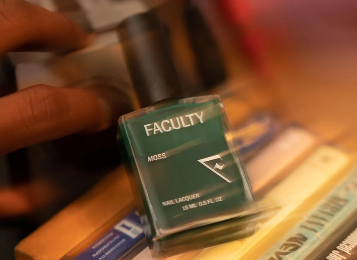 Green nail polish bottle labeled 'FACULTY' with the word 'MOSS' and 'NAIL LACQUER' on the front, in a hand, on a wooden surface with other bottles and magazines.