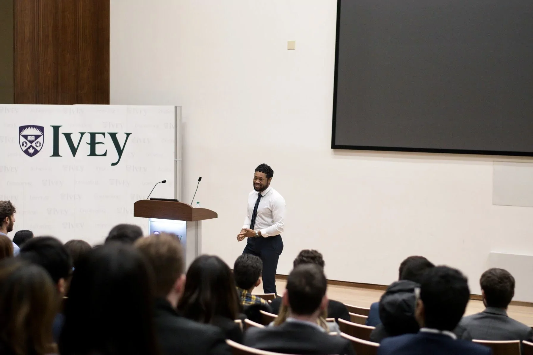 Fenton Jagdeo giving a presentation in a lecture hall with an audience of students, IVEY logo on screen and a large blank monitor on the wall.