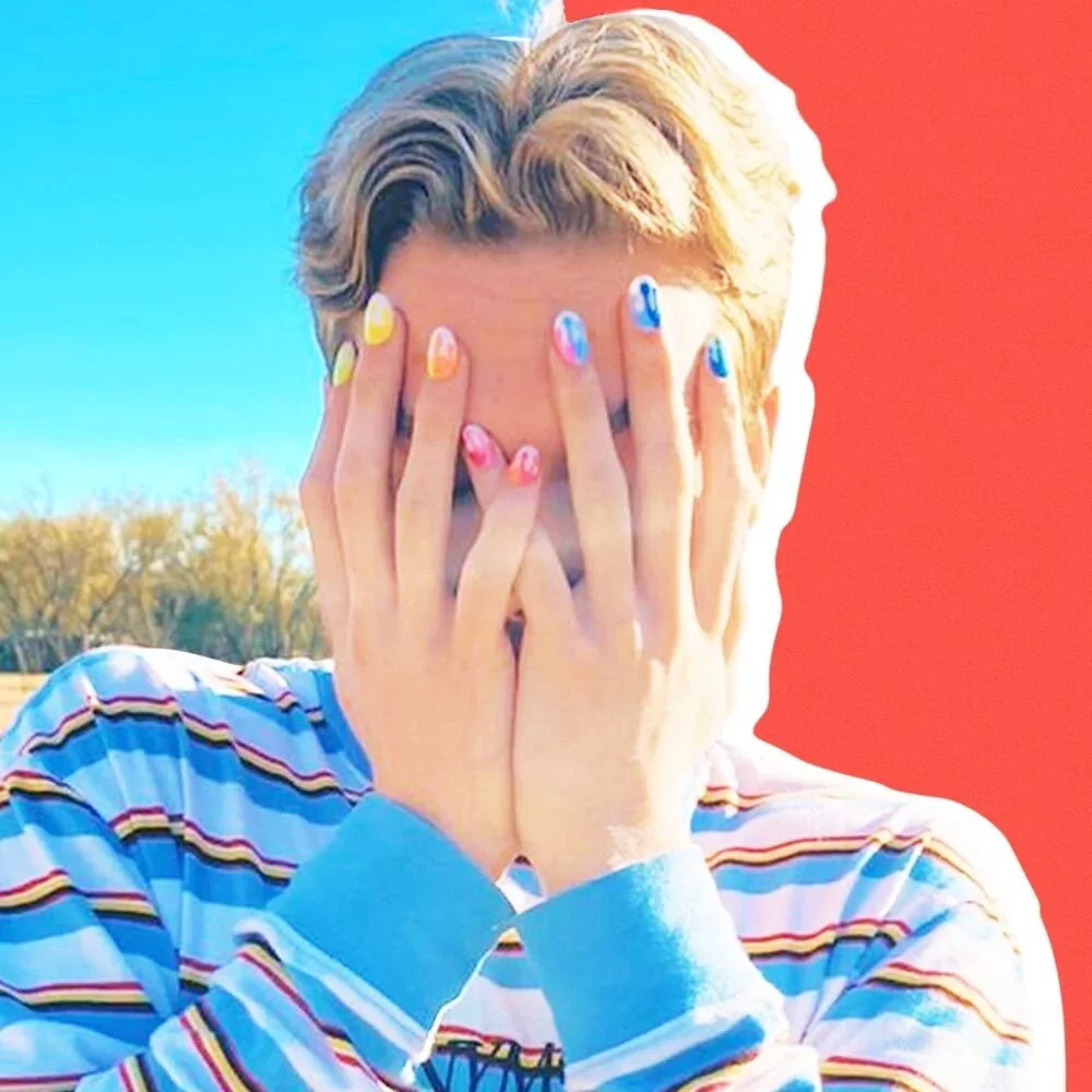 Person with vibrant multicolored painted nails covering their face outdoors, wearing a striped shirt, with a colorful background.