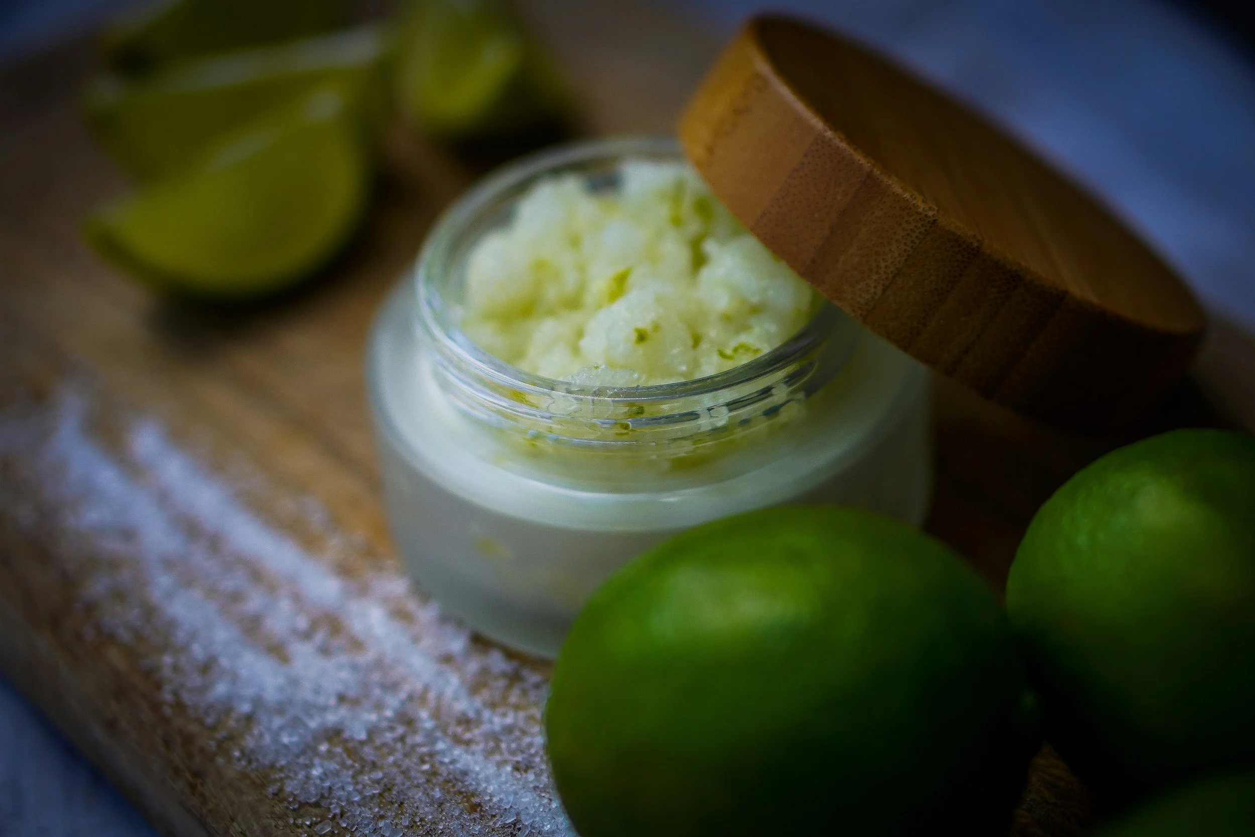 A jar of green body sugar scrub on a wooden surface, surrounded by fresh limes and lime wedges.