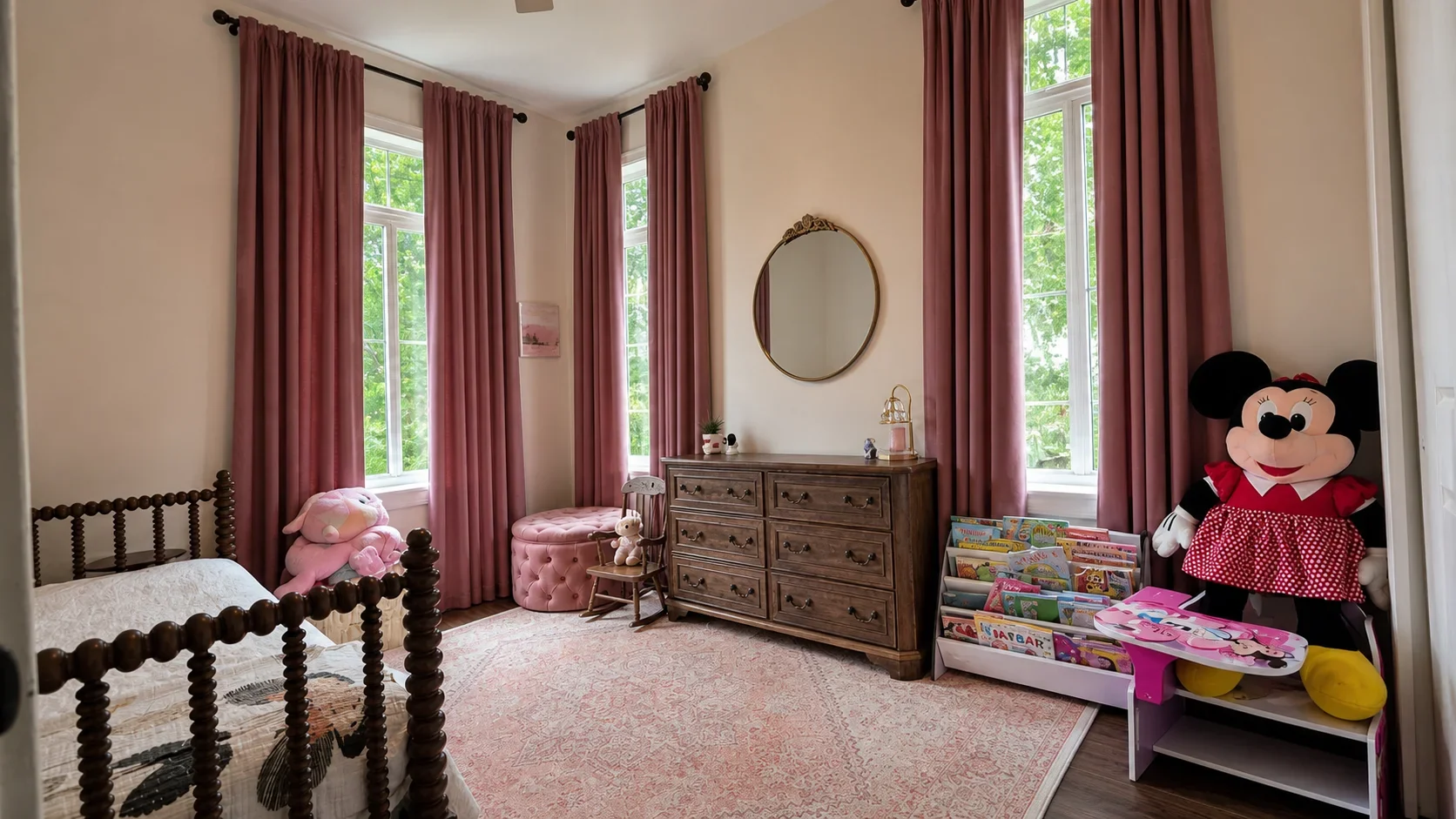 A child's bedroom with large windows, pink curtains, a wooden bed with a stuffed animal, a pink tufted ottoman, a wooden dresser, and shelves filled with children's books and a Minnie Mouse plush doll.