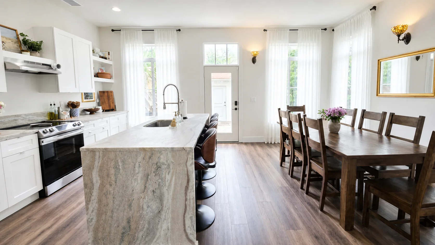 A bright kitchen and dining area with white cabinets, a marble island with a sink and barstools, a wooden dining table with eight chairs, large windows with white curtains, and a mirror on the wall.