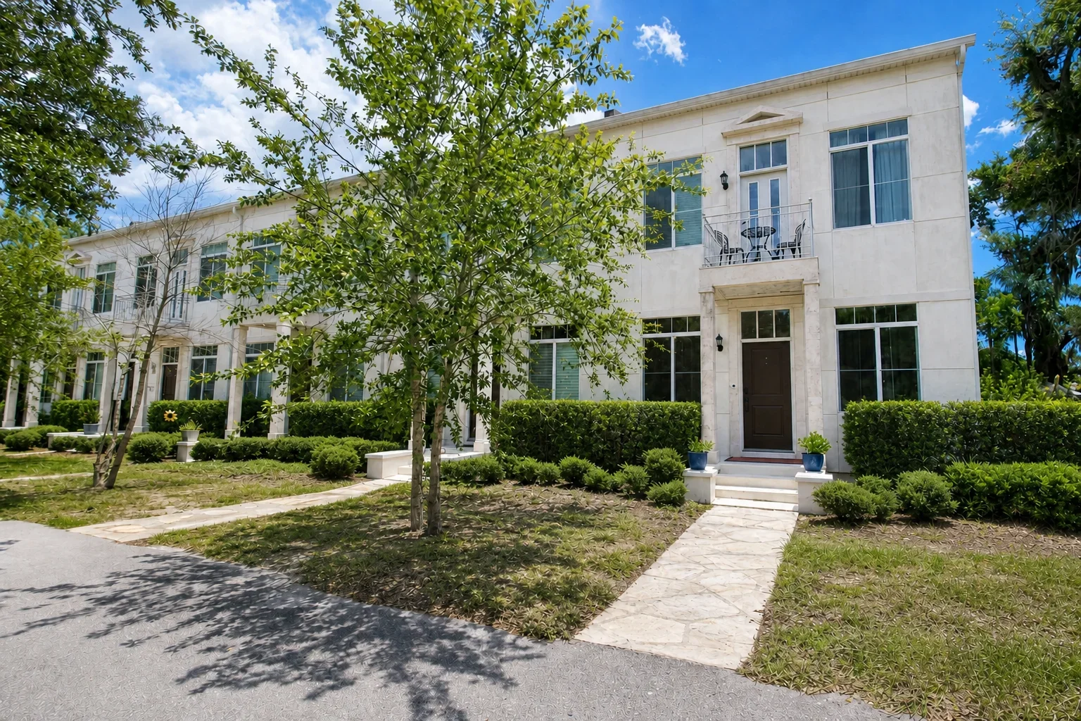 Front view of a modern white two-story residential building with large windows, a small balcony with patio furniture, surrounded by green shrubbery, trees, and a paved walkway under a partly cloudy sky.
