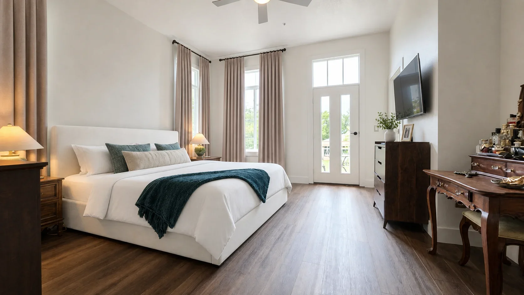 A bright bedroom with a large white bed, beige curtains, a wooden nightstand with a lamp, and a TV mounted on the wall opposite the bed. There is a door with windows leading outside, and a vintage wooden desk with various objects on it.