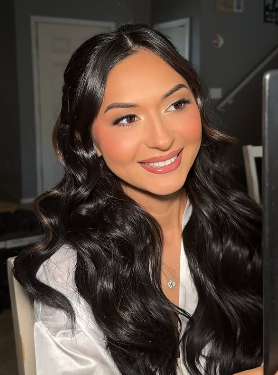 A woman with long, wavy black hair smiling and looking slightly to the side, sitting in a room with a door and staircase in the background.