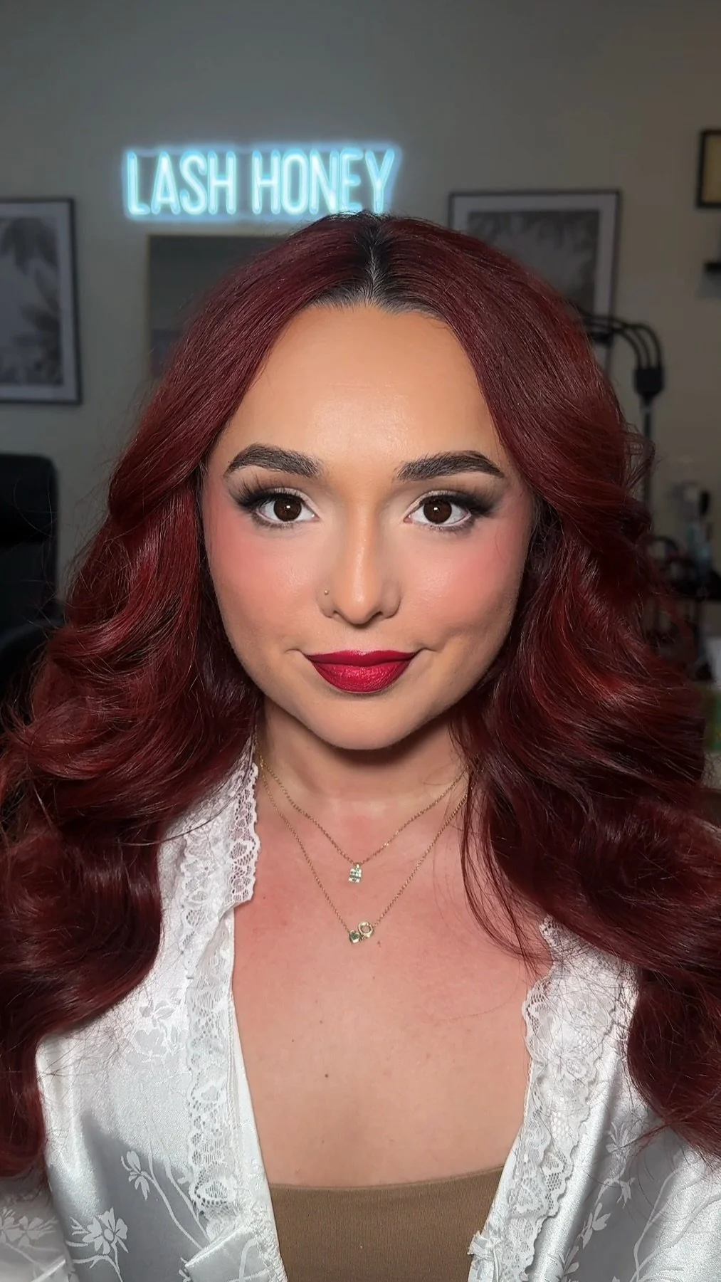 Close-up of a woman with red hair, makeup, and jewelry, in front of a wall with a neon sign that reads 'Lash Honey'.