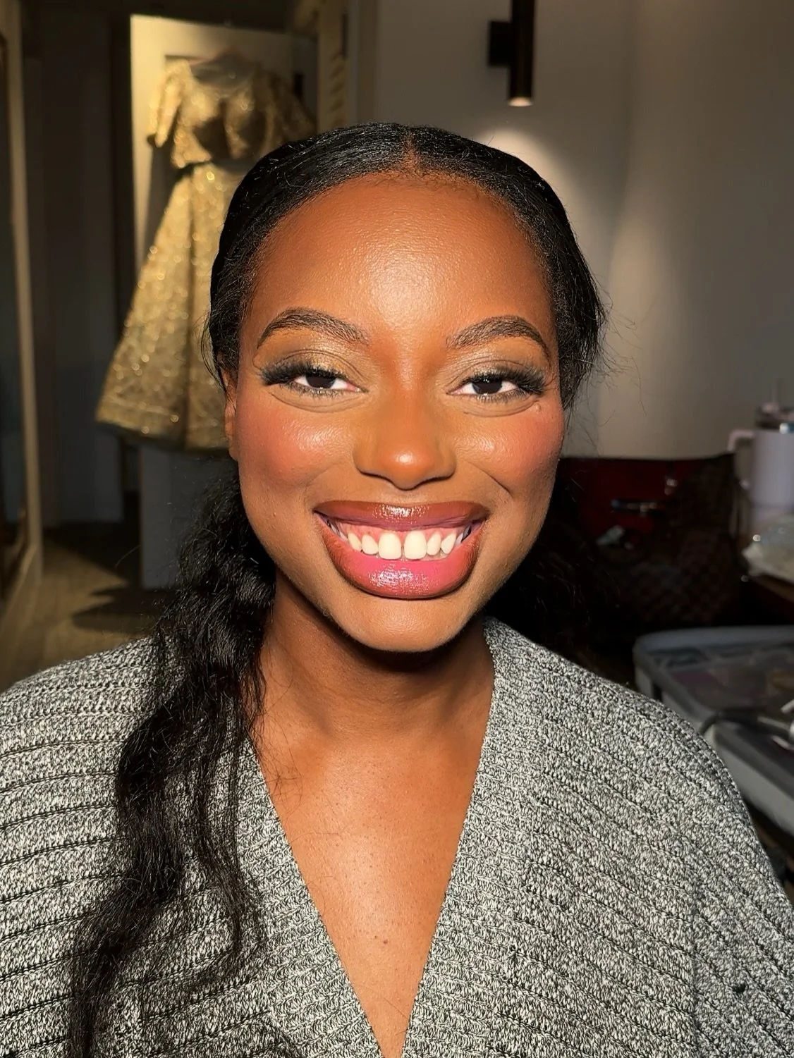 Close-up of a smiling woman with dark hair, makeup, and a grey textured top, in an indoor setting with a gold dress displayed in the background.