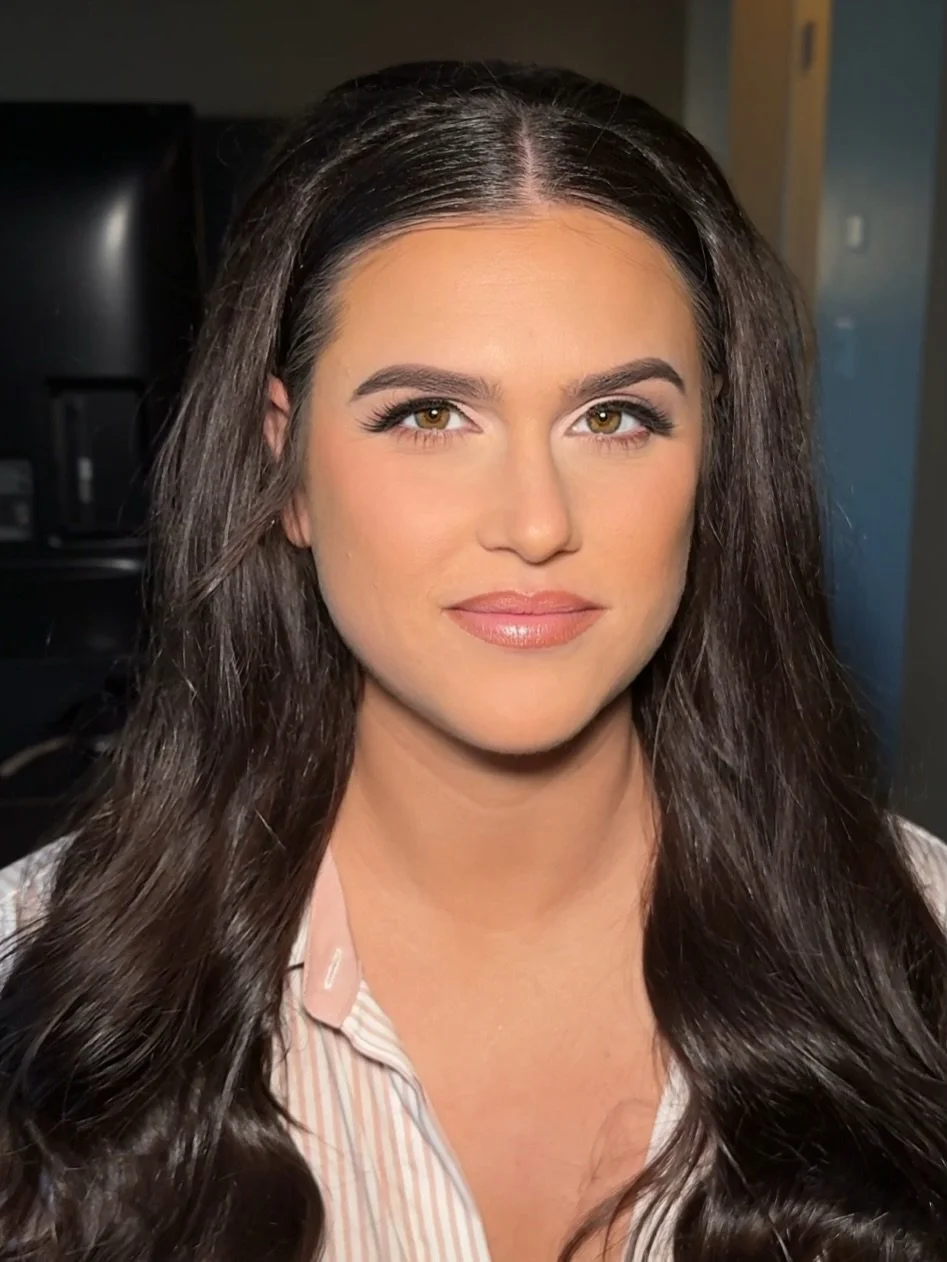 Close-up portrait of a woman with long, dark brown hair, brown eyes, and makeup, smiling slightly, wearing a striped shirt.