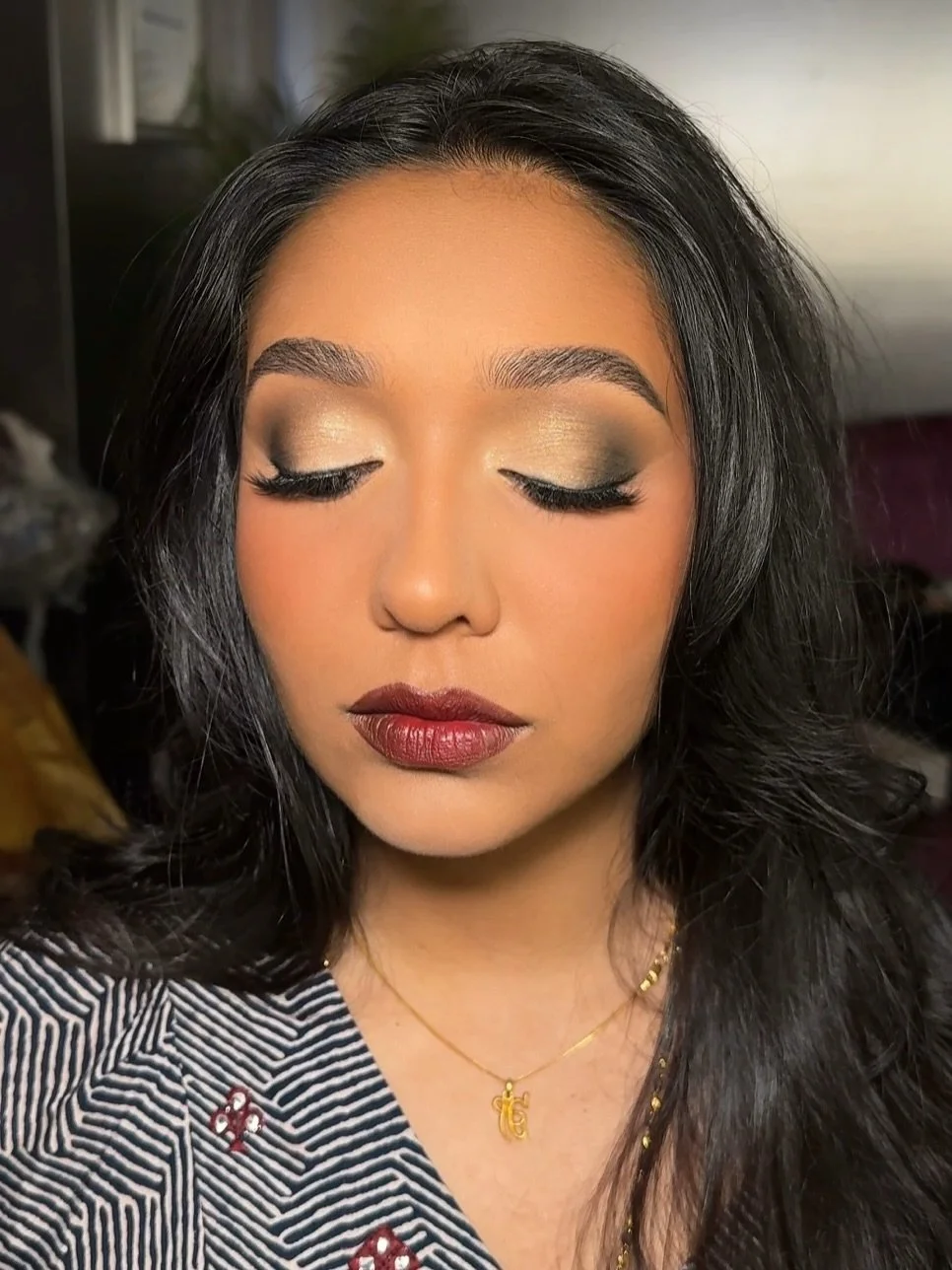 Portrait of a woman with closed eyes, wearing makeup, gold jewelry, and a patterned top.