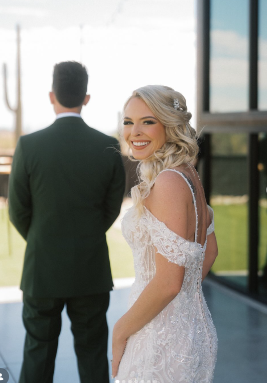 A bride with long blonde hair in a lace wedding dress smiling at the camera, with a groom in a dark suit standing with his back facing the camera in the background.