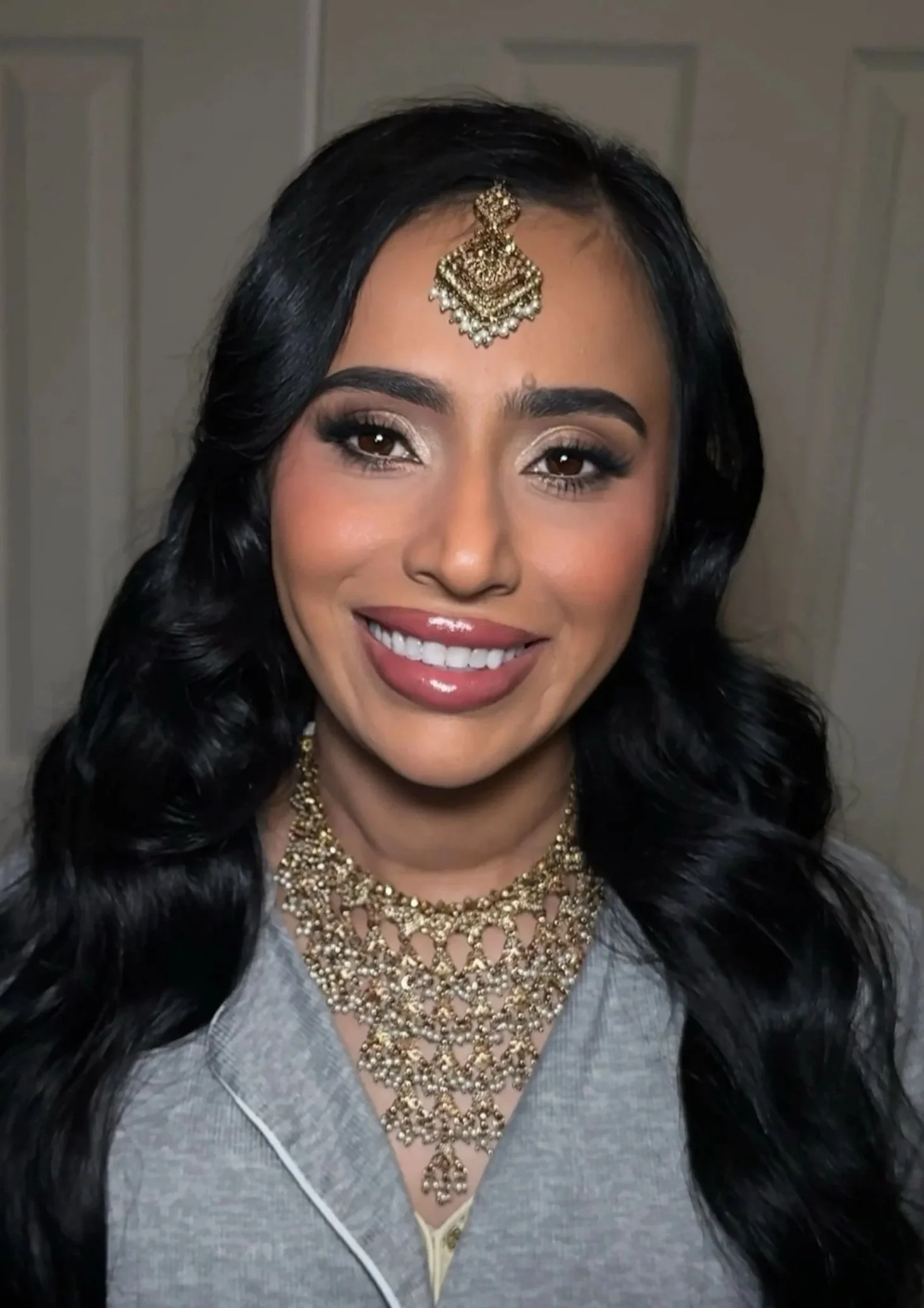 A woman with long black wavy hair wearing traditional gold jewelry, including a maang tikka, necklace, and earrings, smiling at the camera.