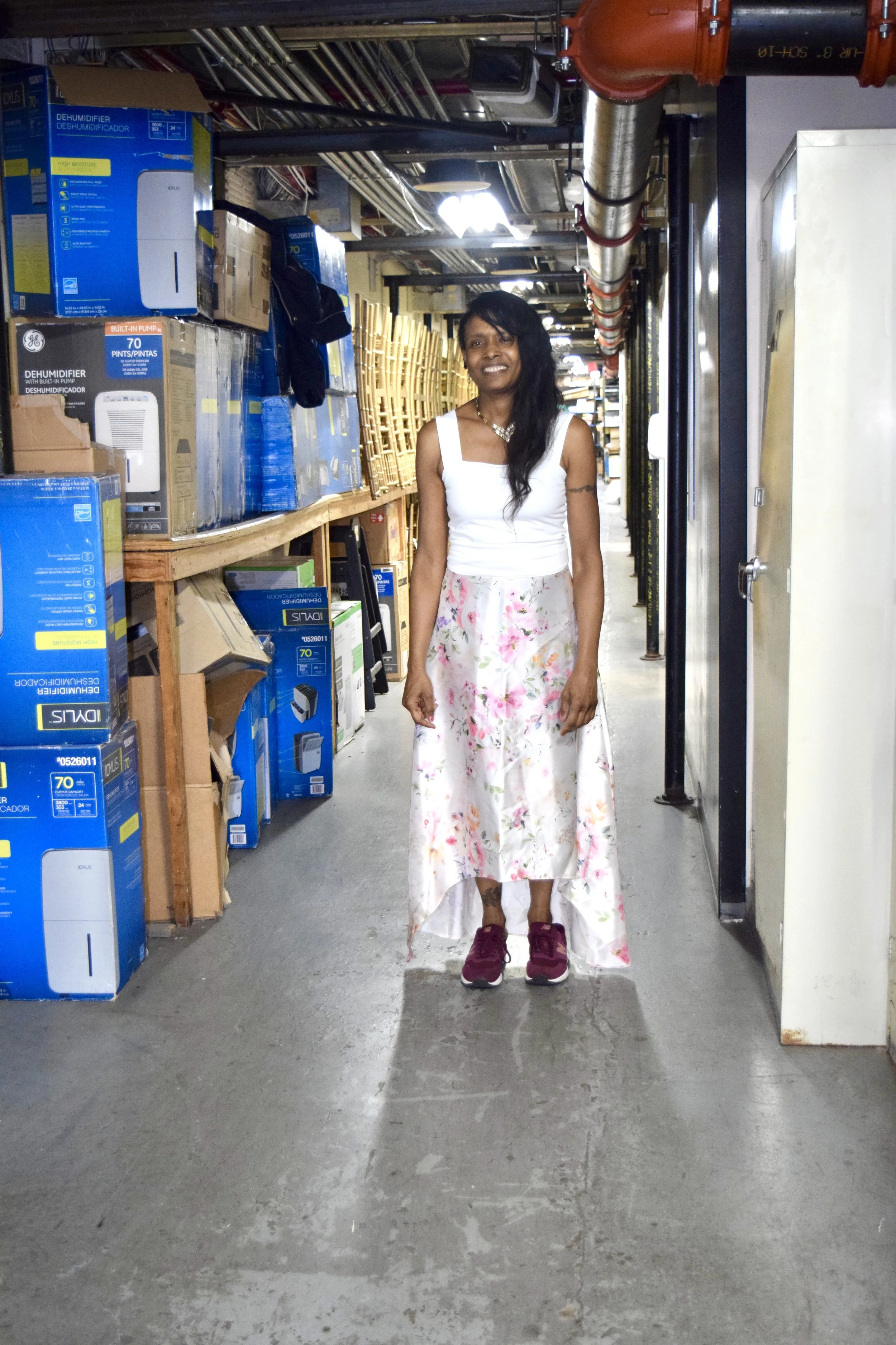 A woman standing in a warehouse aisle, wearing a white sleeveless top, floral skirt, and maroon sneakers, smiling at the camera.