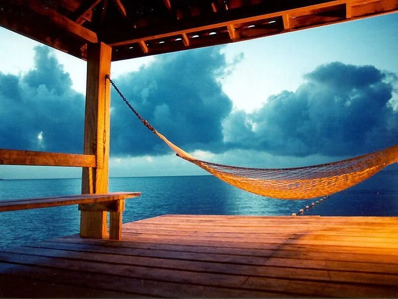 View from a wooden dock with a hammock hanging, overlooking the ocean and cloudy sky during dusk or dawn.