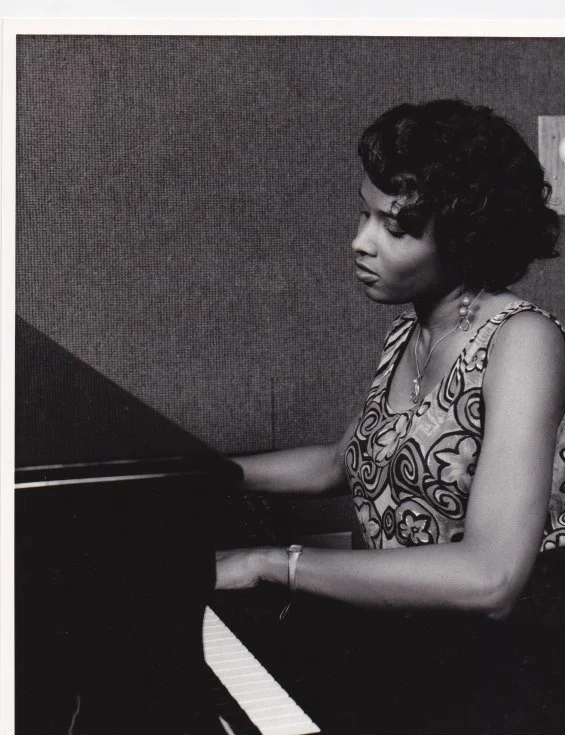 A woman playing a piano, wearing a patterned dress and jewelry, in a black-and-white photograph.