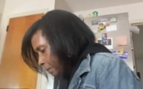 A woman with black hair looking down, wearing a denim jacket, in a kitchen with a refrigerator covered in magnets and notes.