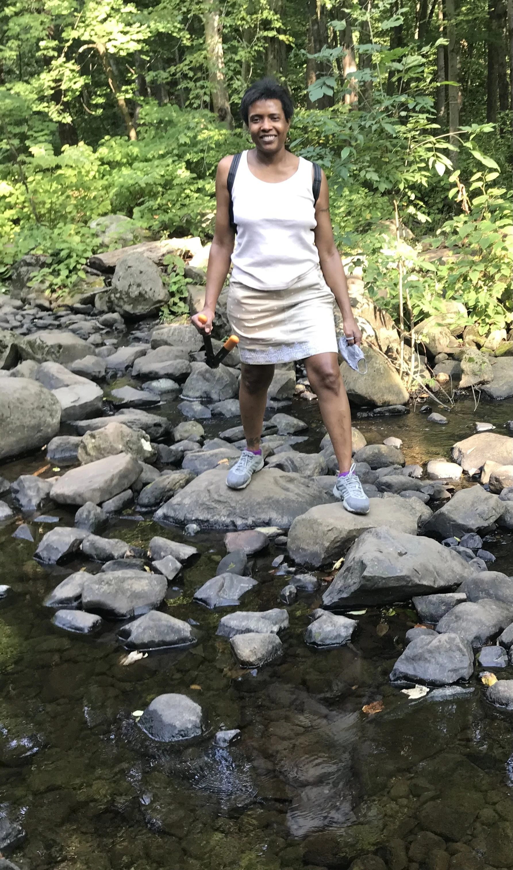 A woman smiling while standing on rocks in a creek in a wooded area, holding a hiking pole in her right hand and a face mask in her left hand.