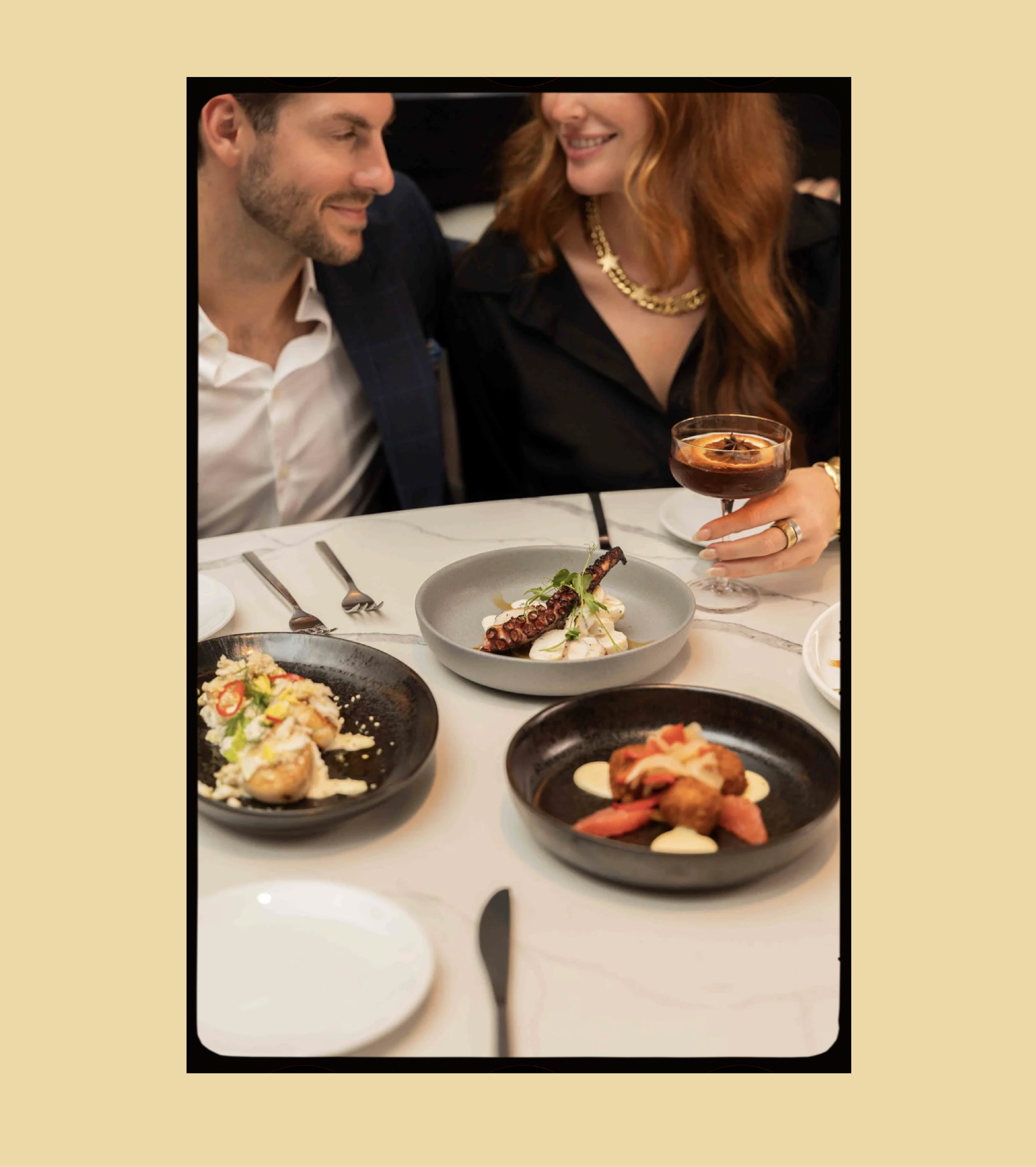 A man and woman smiling at each other at a dinner table with three plated dishes and a cocktail.