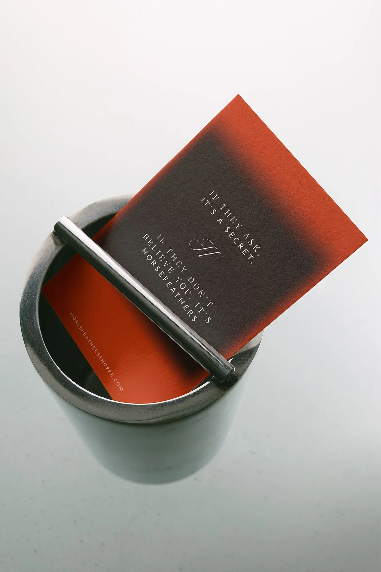 A promotional card inside a cylindrical metal container with a black and red design, featuring a quote about trust and belief, with the website horsefeathershoppe.com printed at the bottom.