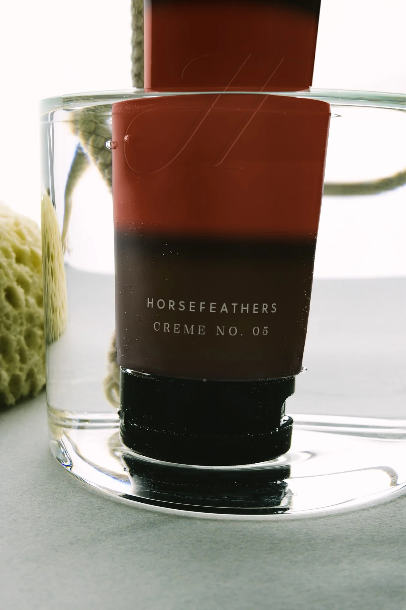 A tube of Horsefeathers Creme No. 05 skincare product partially submerged in water with a glass of water and a yellow sponge to the side.