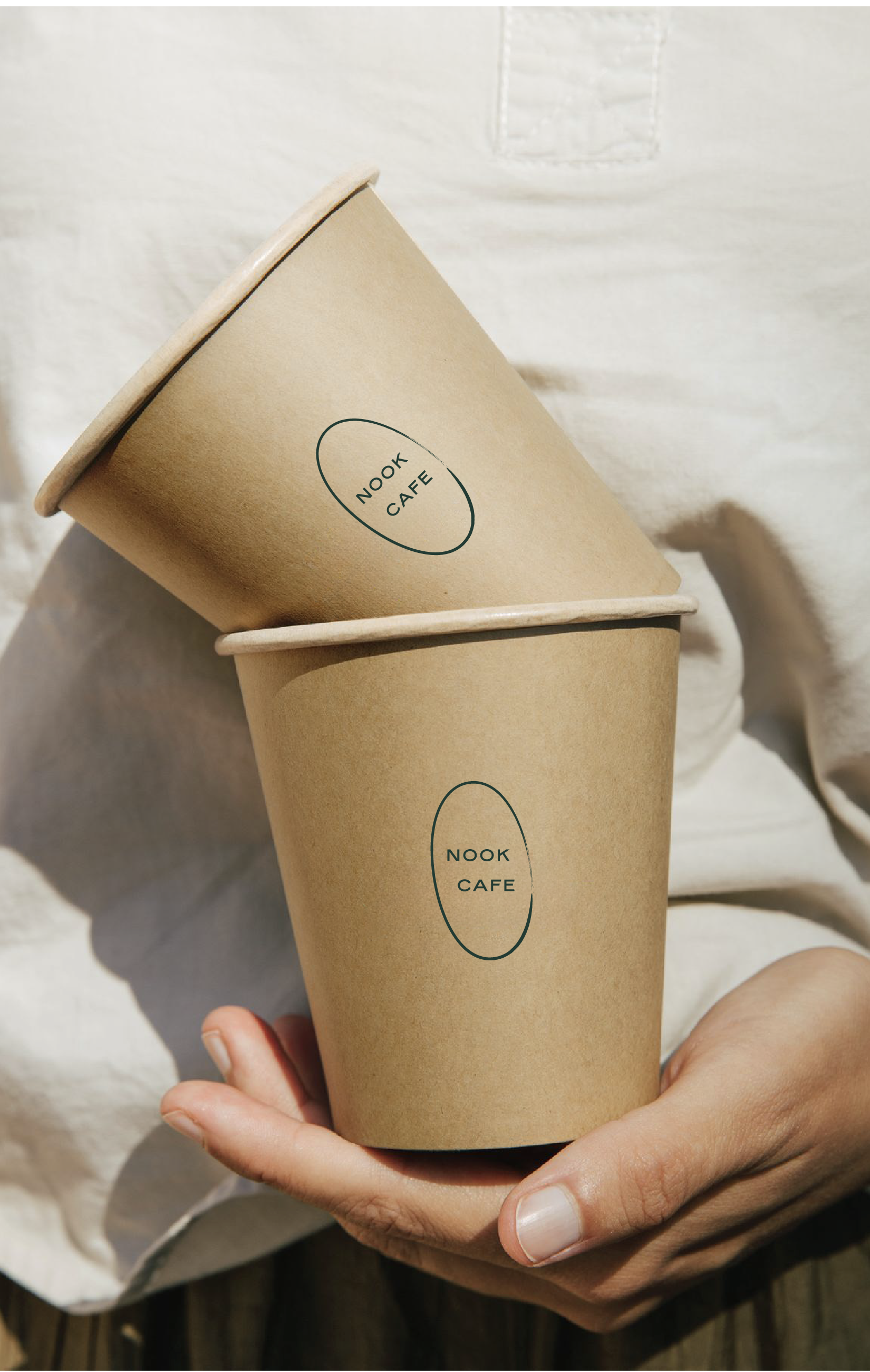 Person holding two brown paper cups with 'Nook Cafe' printed on them, one slightly tilted.