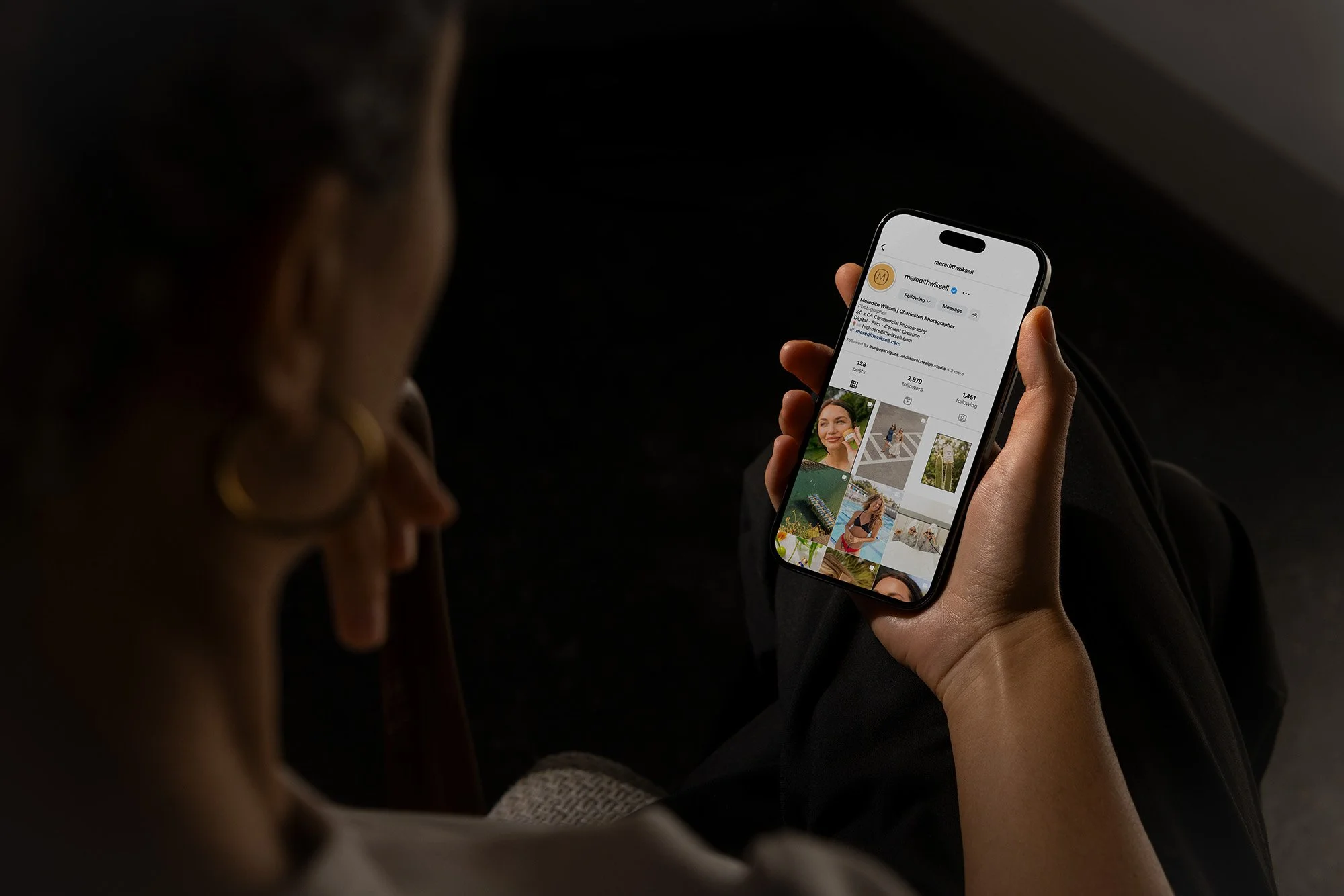 A person is holding a smartphone displaying an Instagram profile with photos of a woman outdoors, including landscapes, a portrait, and a woman in a swimsuit.