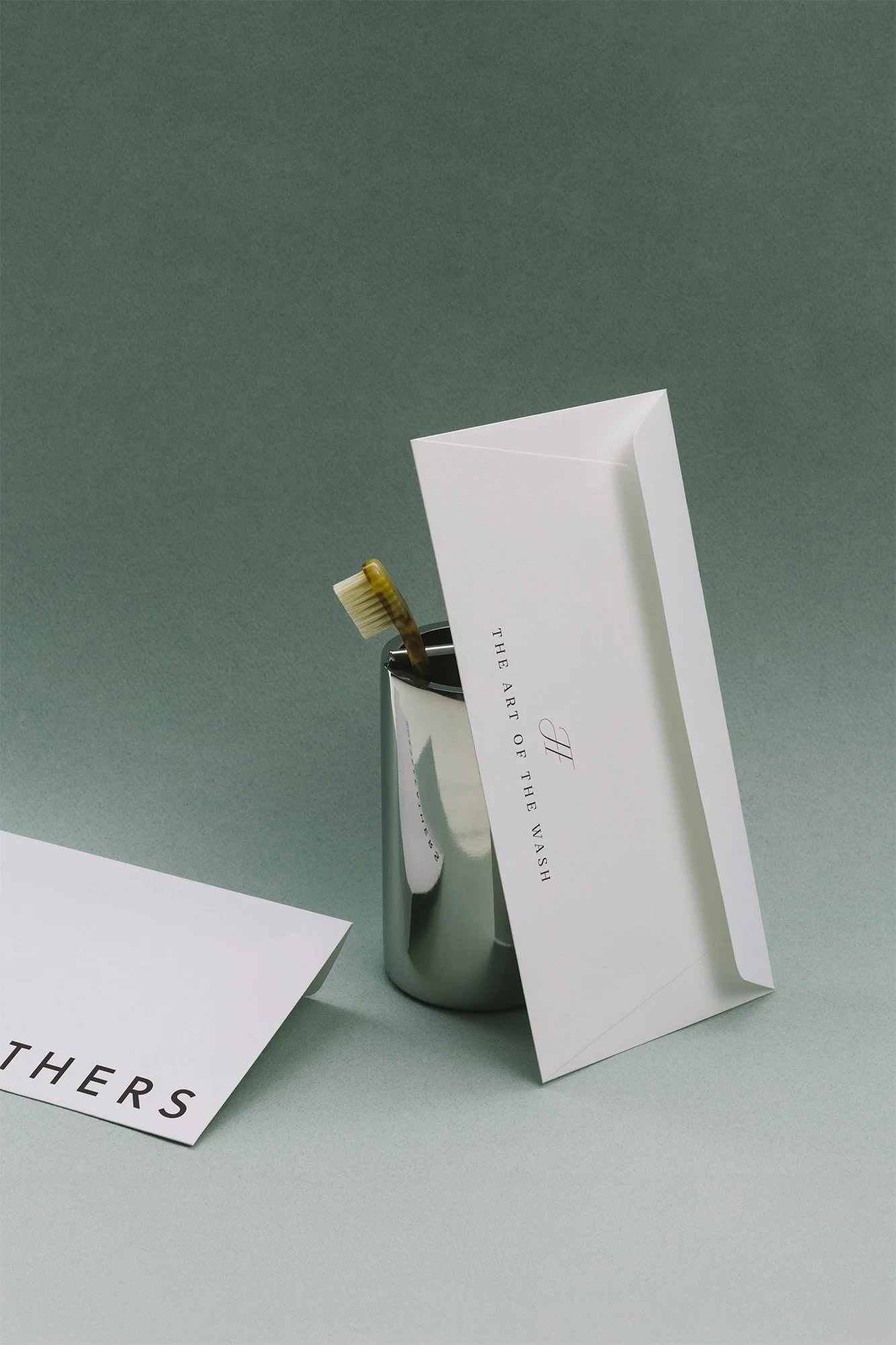 A toothbrush in a silver metal cup, an envelope, and a paper with the quote "The art of the wash" on a green surface.